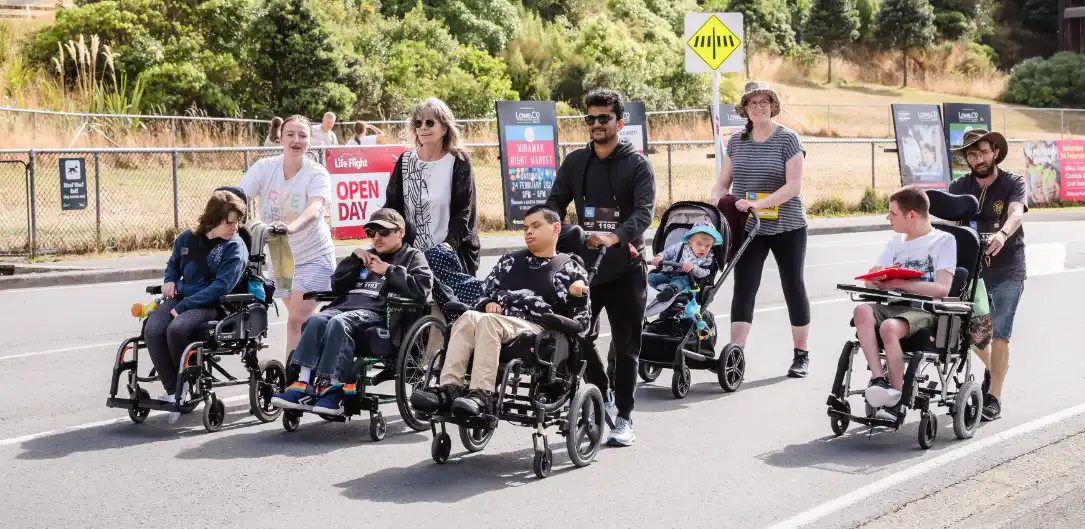 A group of people, including children and adults, are on a walk. Some of them are using wheelchairs. They are outdoors on a paved pathway with greenery and trees in the background. Everyone appears to be engaged and enjoying the walk.