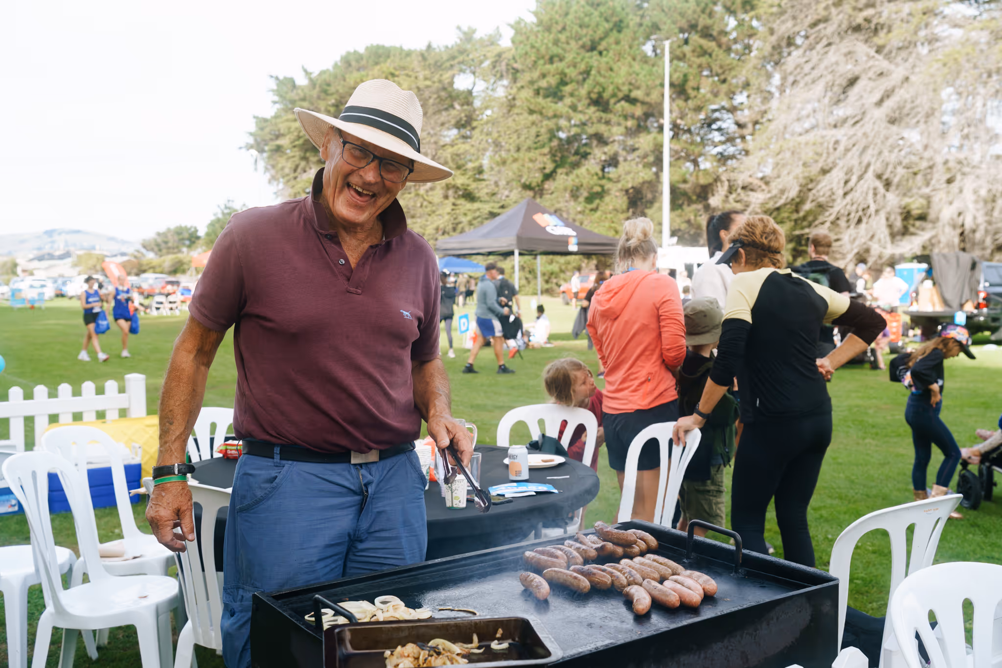 Man in a hat grilling sausages and onions at an outdoor event with people and trees in the background.