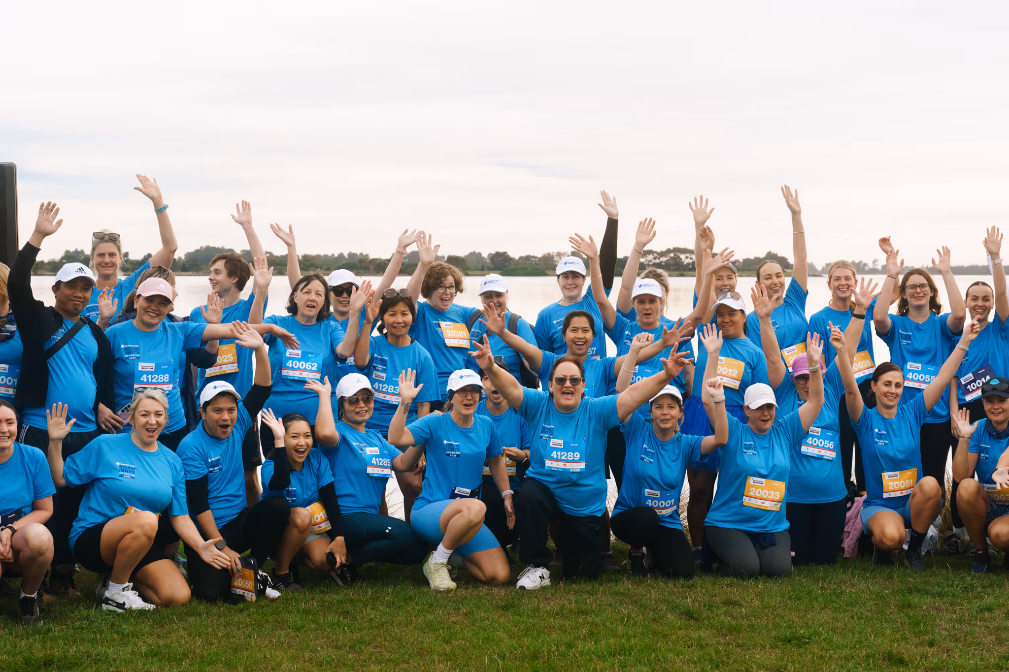 Group of people in blue event shirts raising hands outdoors on grass with water and trees in background.