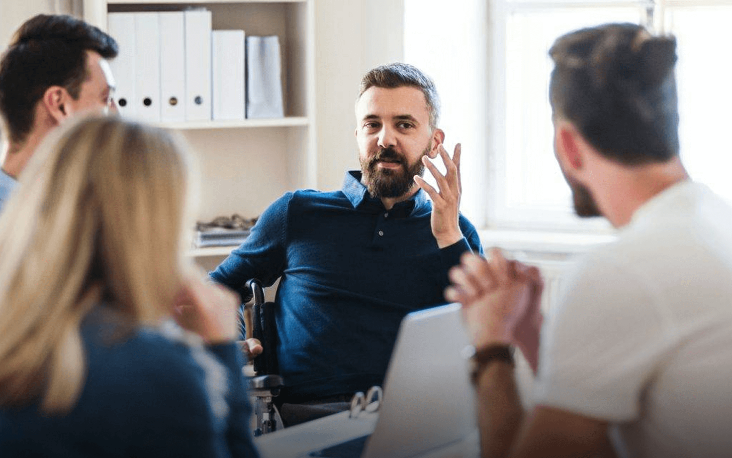 Man in a chair discussing with three colleagues in a bright office setting.