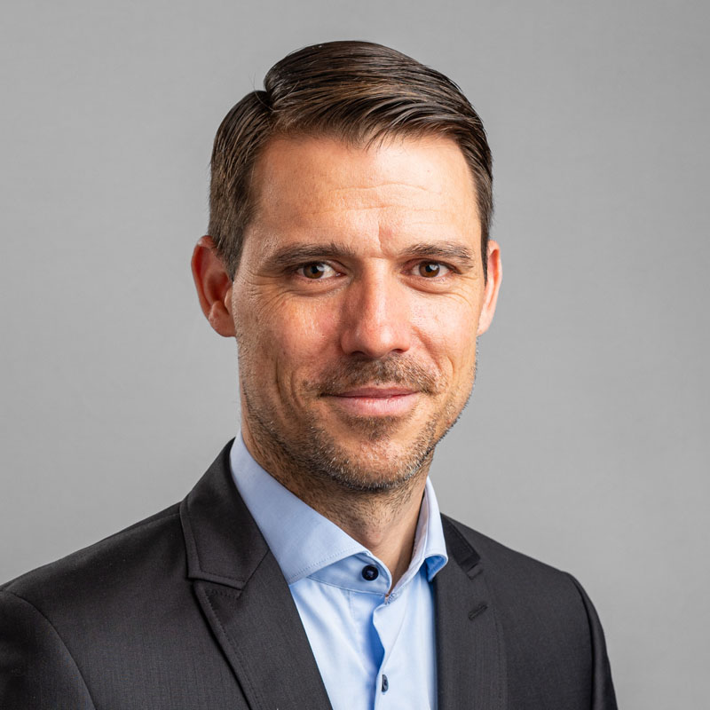 Portrait eines Mannes mit kurzen dunklen Haaren in einem dunklen Anzug und hellblauem Hemd vor grauem Hintergrund.