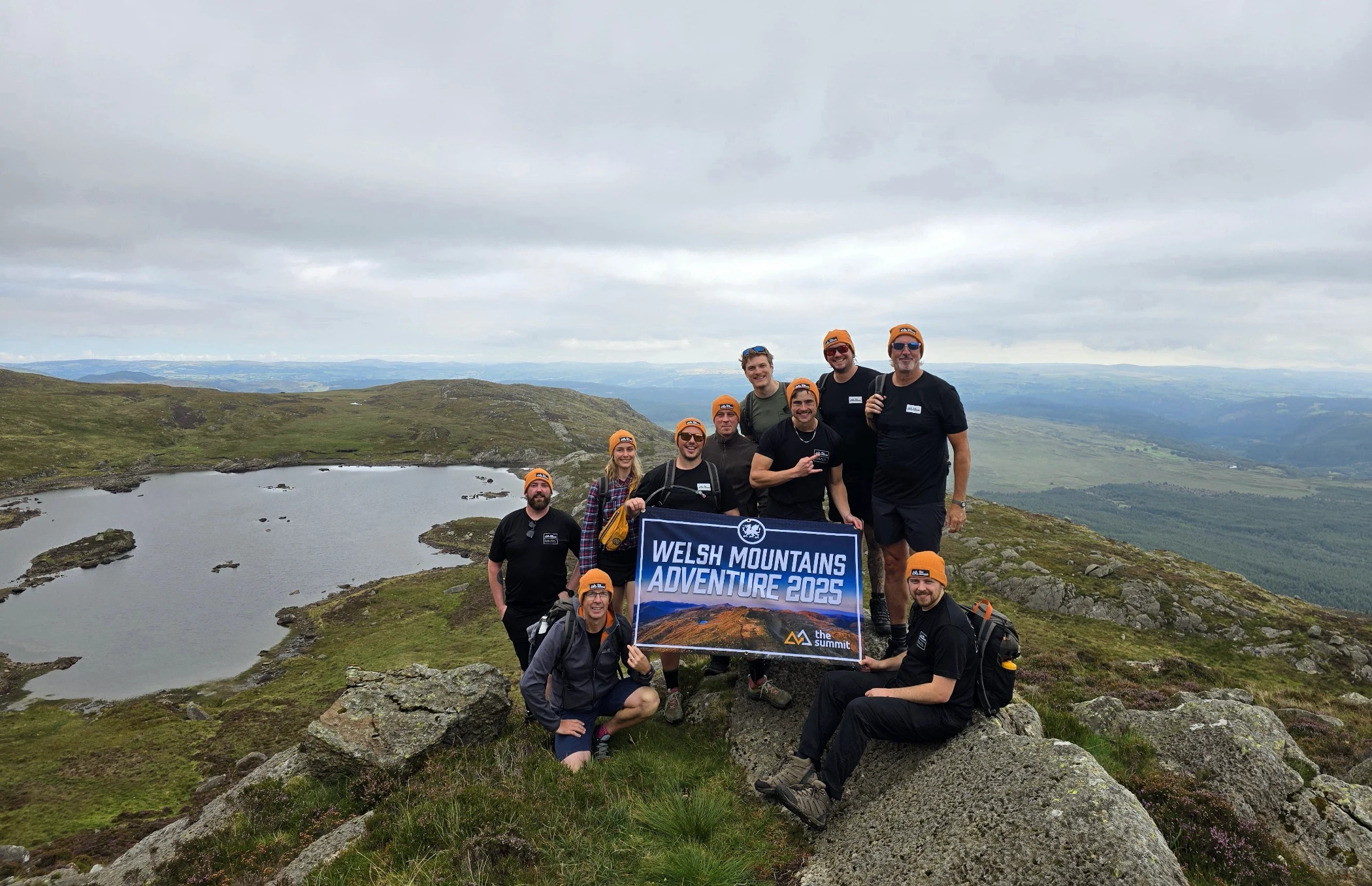 Group of nine hikers wearing matching orange beanies posing with a 'Welsh Mountains Adventure 2025' banner on a rocky hill overlooking a lake and rolling mountains under a cloudy sky.