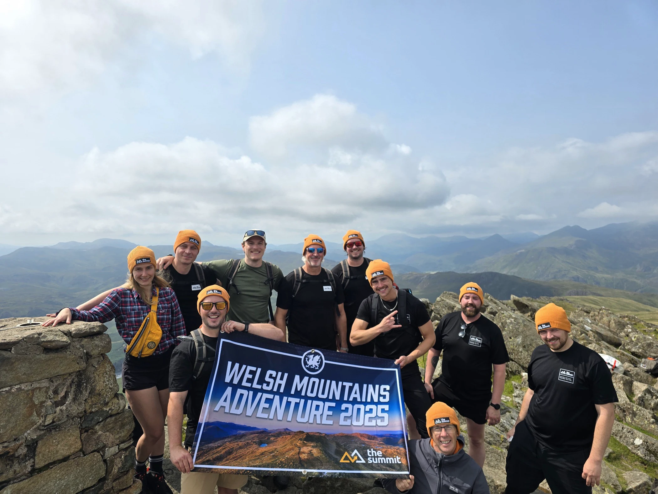 Group of hikers wearing orange beanies holding a banner that reads 'Welsh Mountains Adventure 2025' with mountainous landscape in the background.