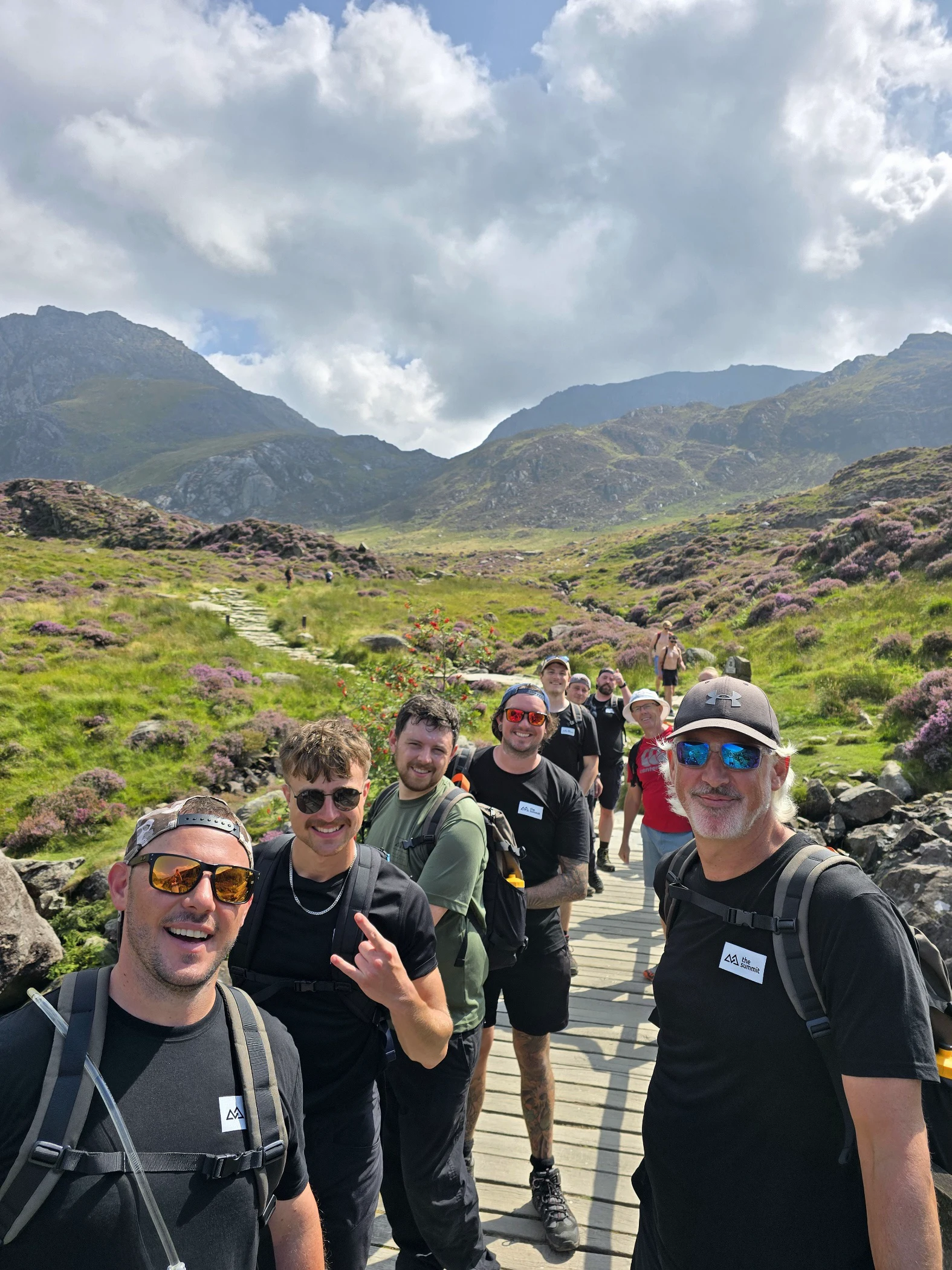 Group of smiling hikers standing on a wooden bridge in a green mountainous landscape under a partly cloudy sky.