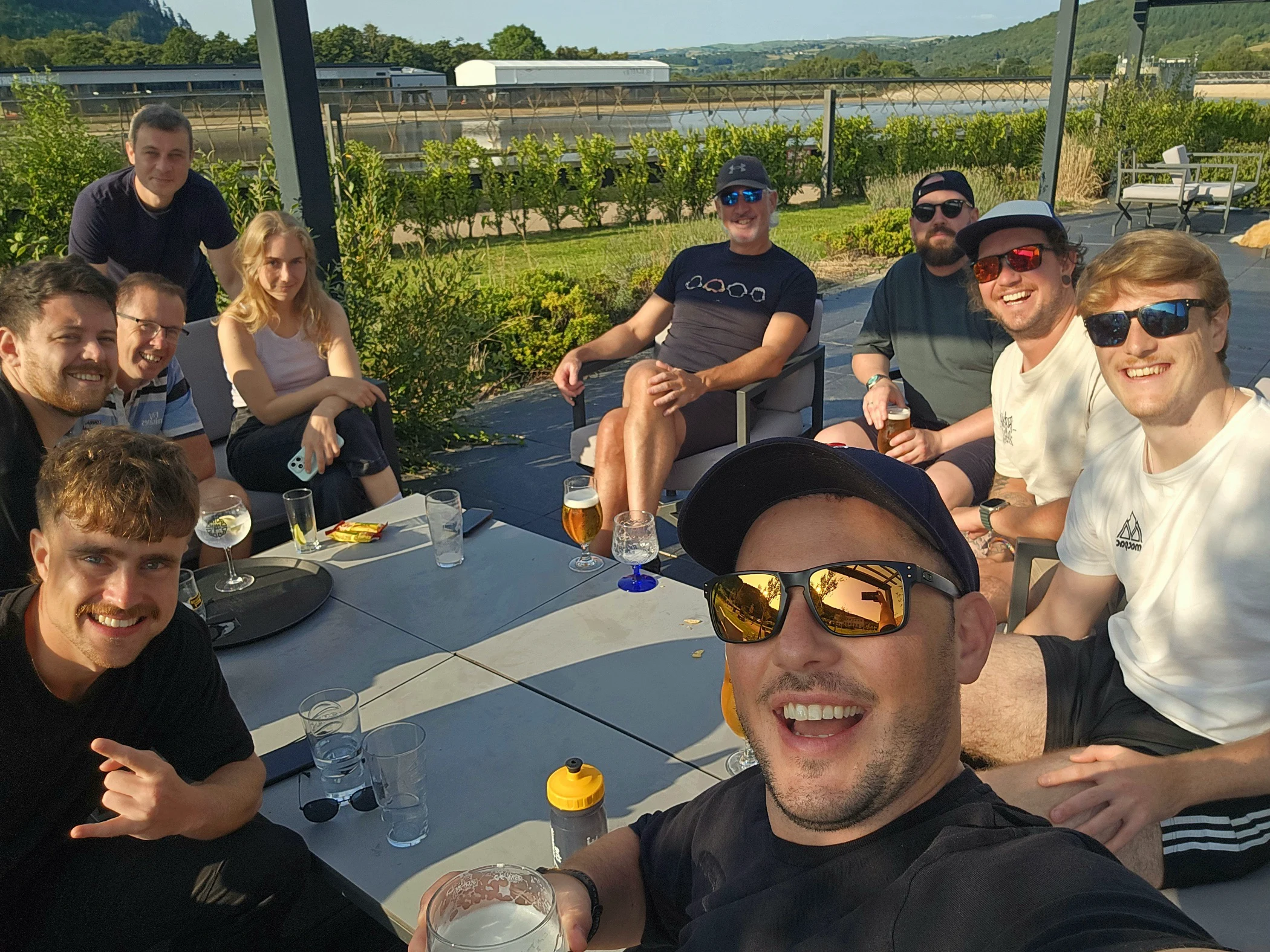 Group of ten people outdoors sitting around tables with drinks, smiling and enjoying sunny weather.