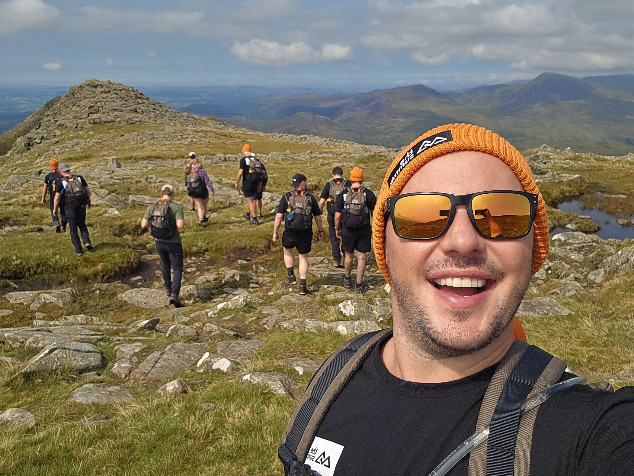 Smiling man wearing an orange beanie and sunglasses taking a selfie while hiking with a group on rocky terrain with mountains in the background.