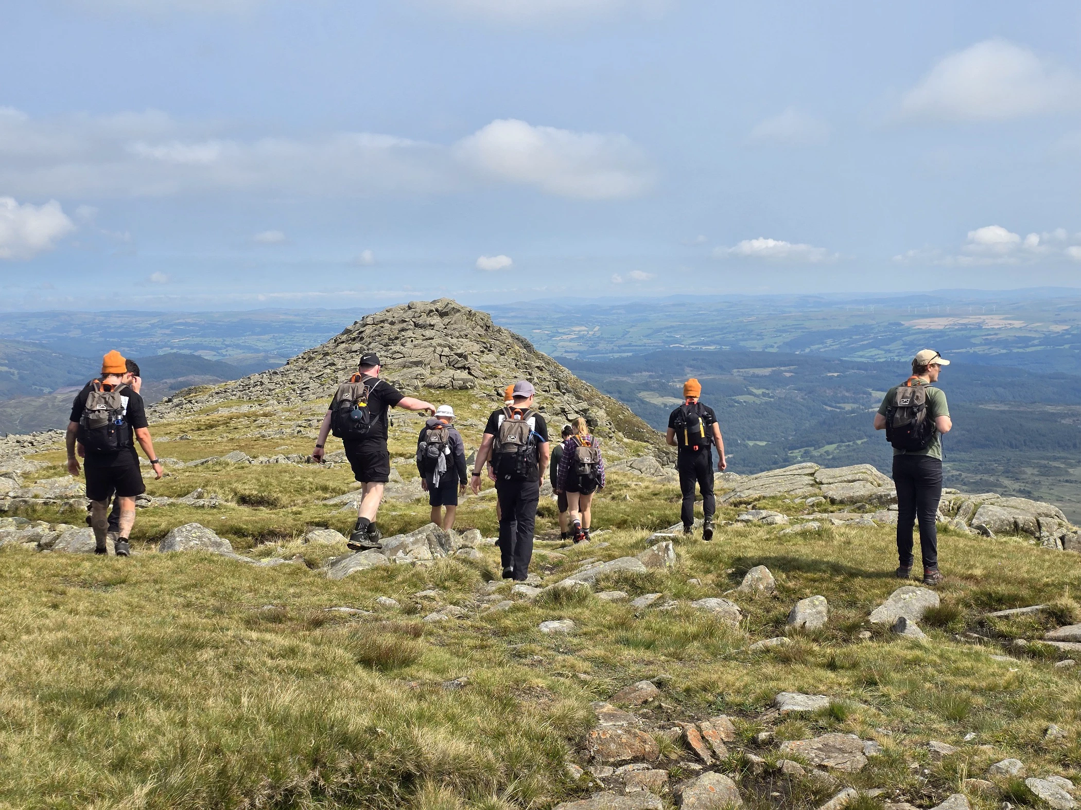 Group of hikers walking along a grassy and rocky mountain ridge with expansive valley views under a partly cloudy sky.