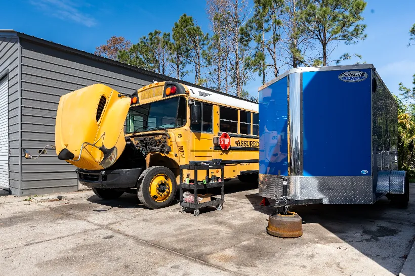 School bus repairs underway with hood raised, exposing engine; tool cart positioned beside front wheel.
