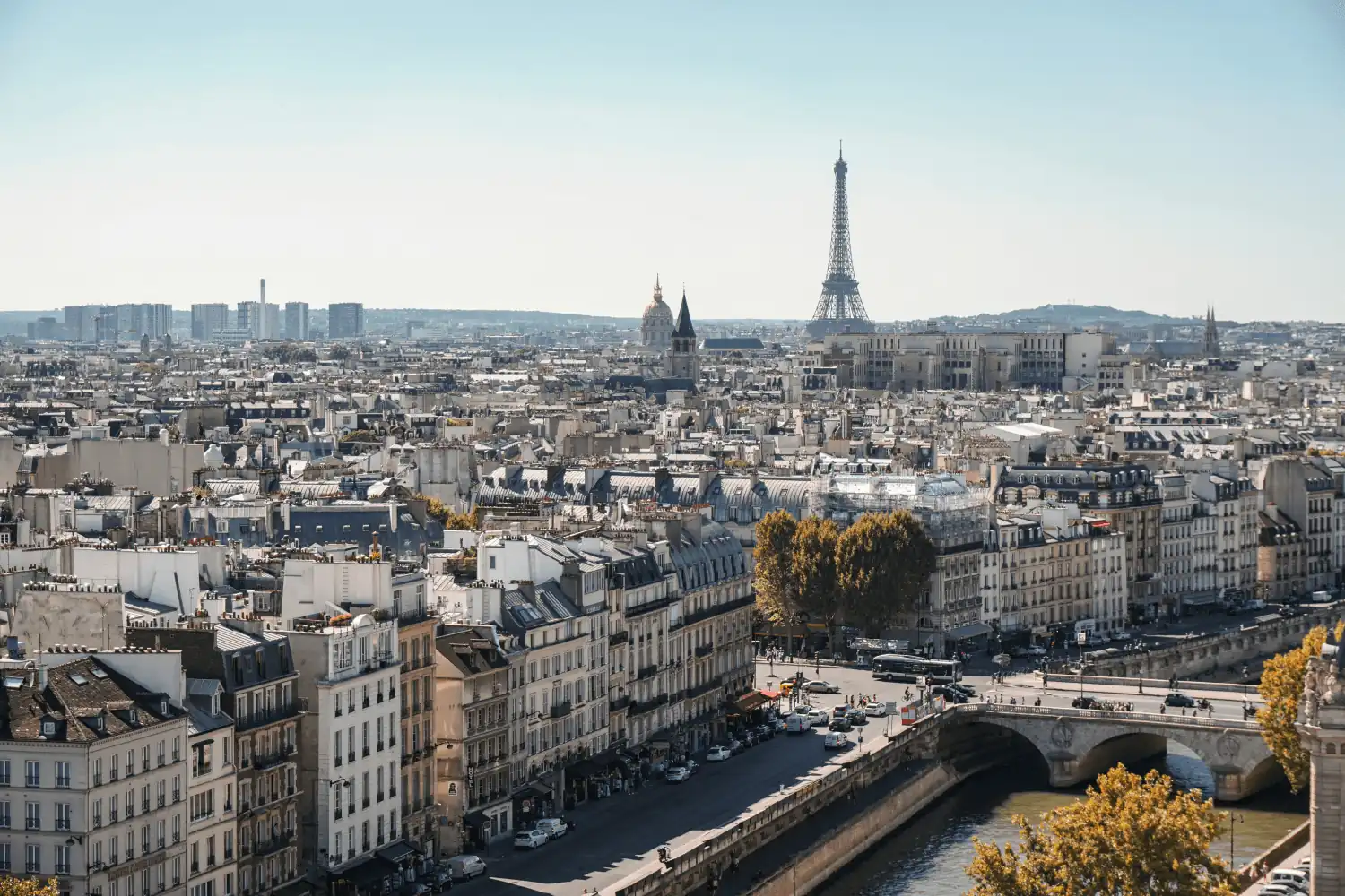 Vue panoramique de la ville de Paris avec la tour Eiffel en arrière-plan, des bâtiments haussmanniens au premier plan, et la Seine traversée par un pont.
