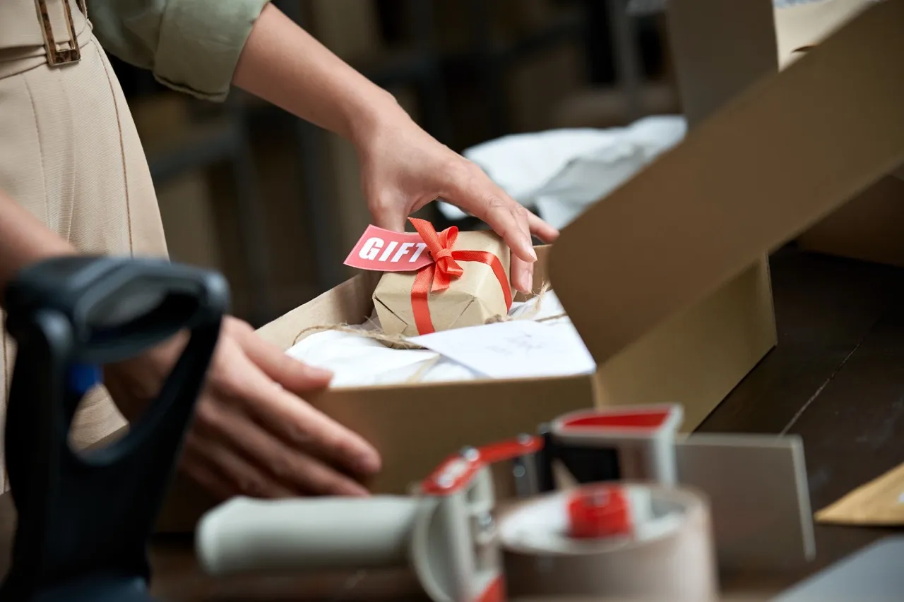 A hand holding gift wrapped box with a label saying "gift" and putting it into a larger box.