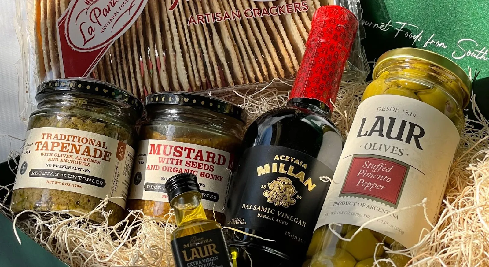A food gift box filled with jars that contain tapenade, mustard, balsamic vinegar, olives, and crackers