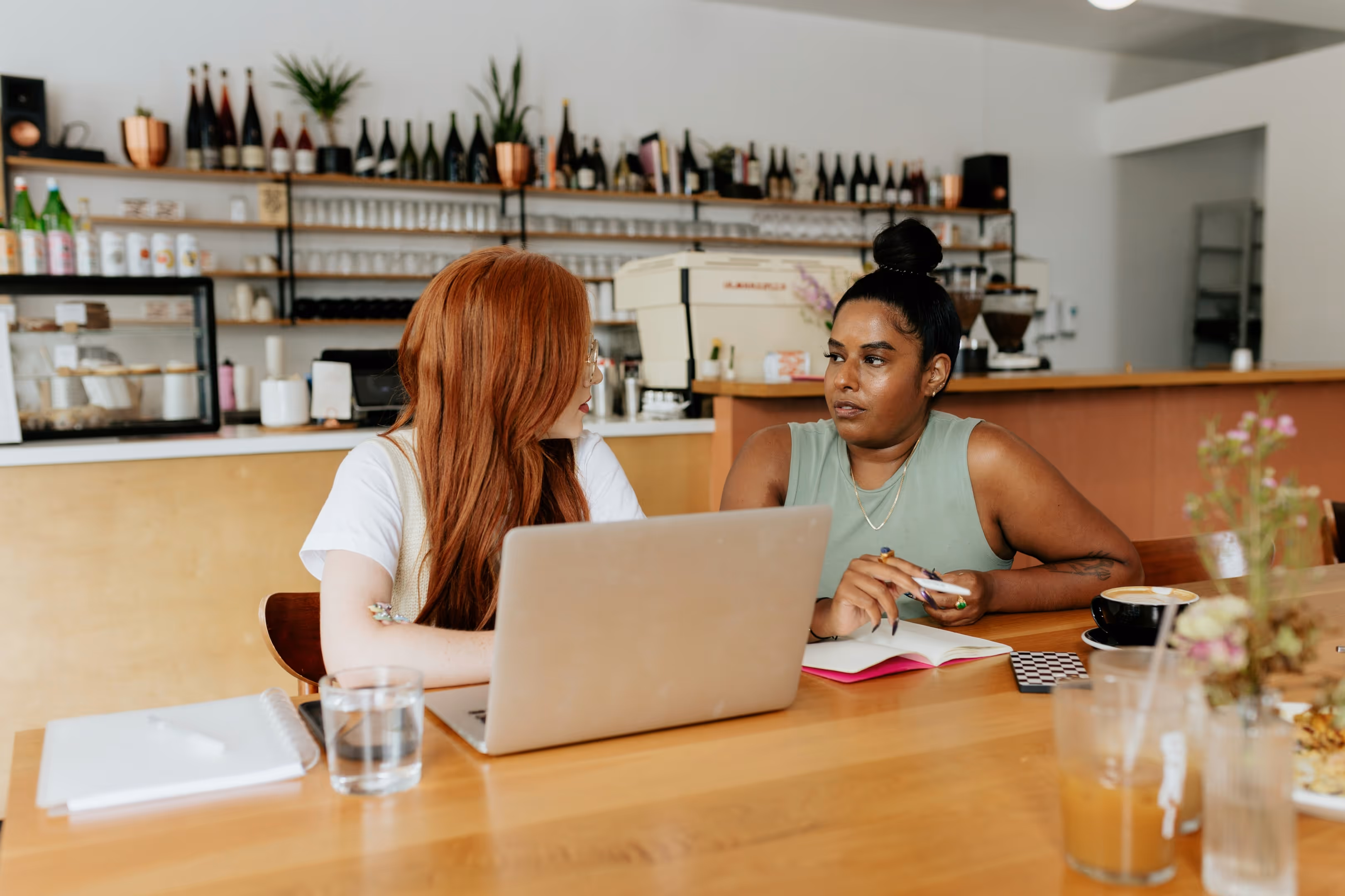 Two women chatting at a coffee shop about business; one training the other on how to run her businesses logistics effectively