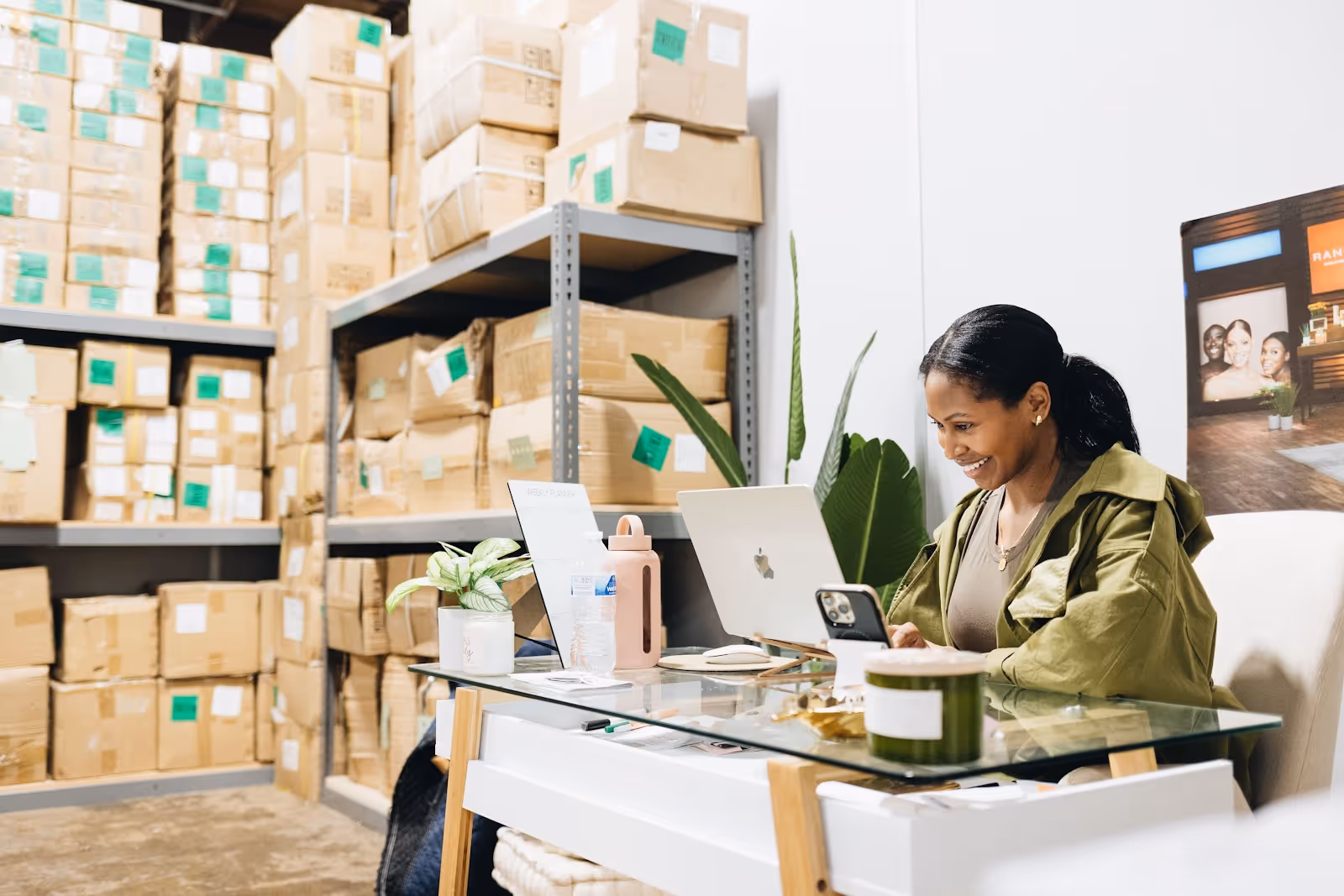 A woman inside a co-warehousing facility, looking at her computer smiling, surrounded by organized boxes on shelves
