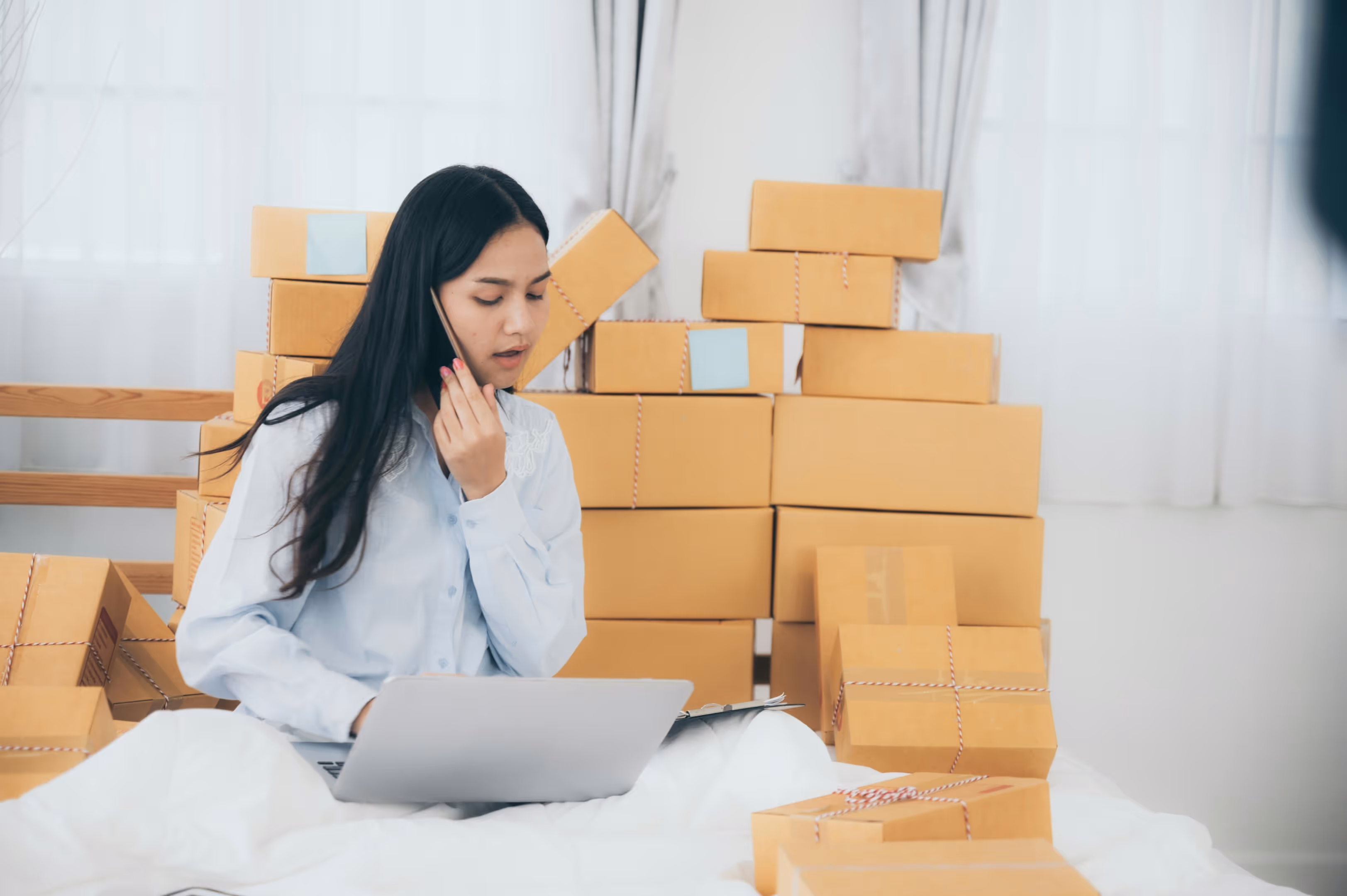 A woman sitting on the floor surrounded by boxes while talking on the phone and looking at a laptop