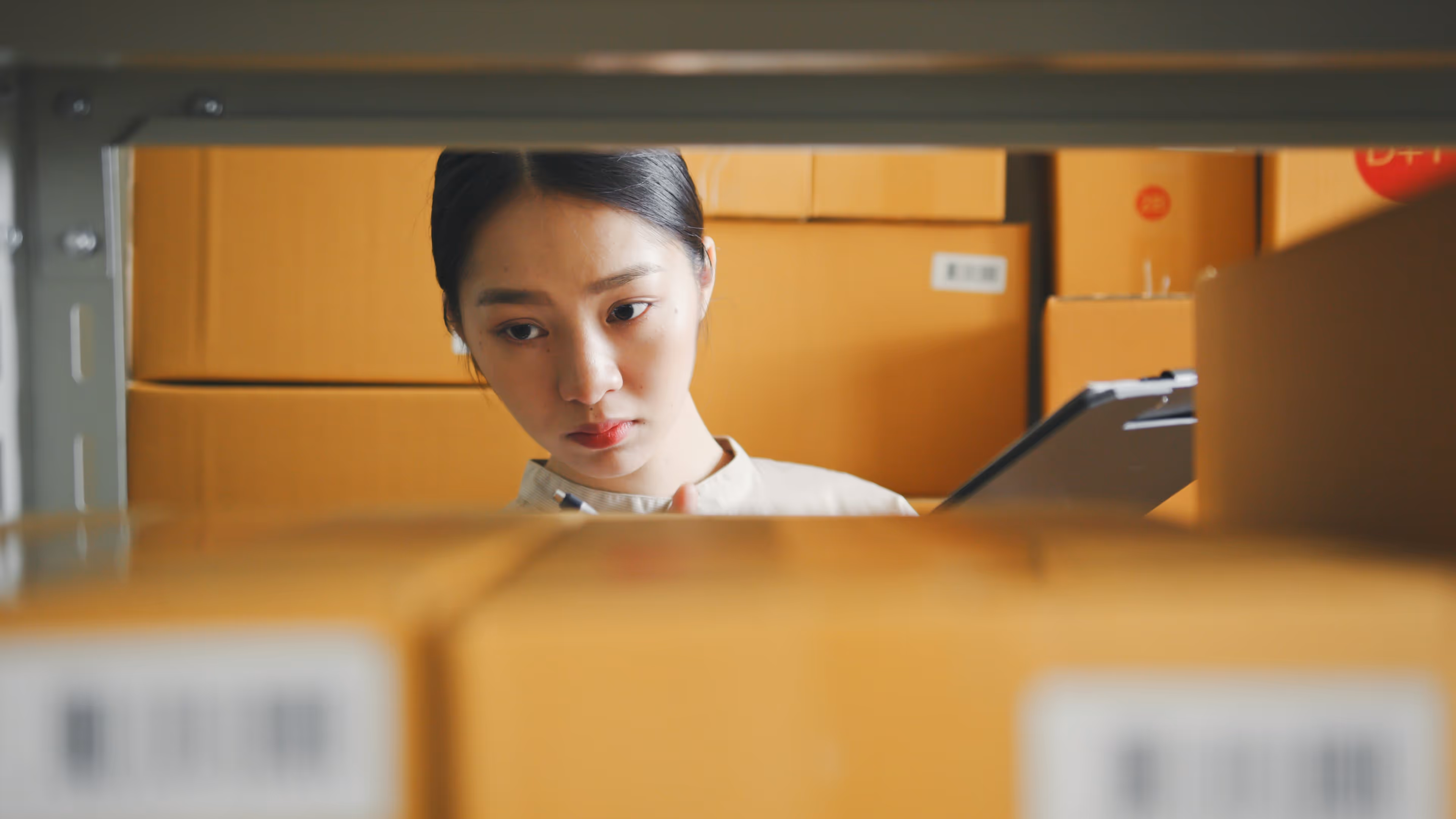 A woman peeking through a shelf filled with boxes while holding a clipboard