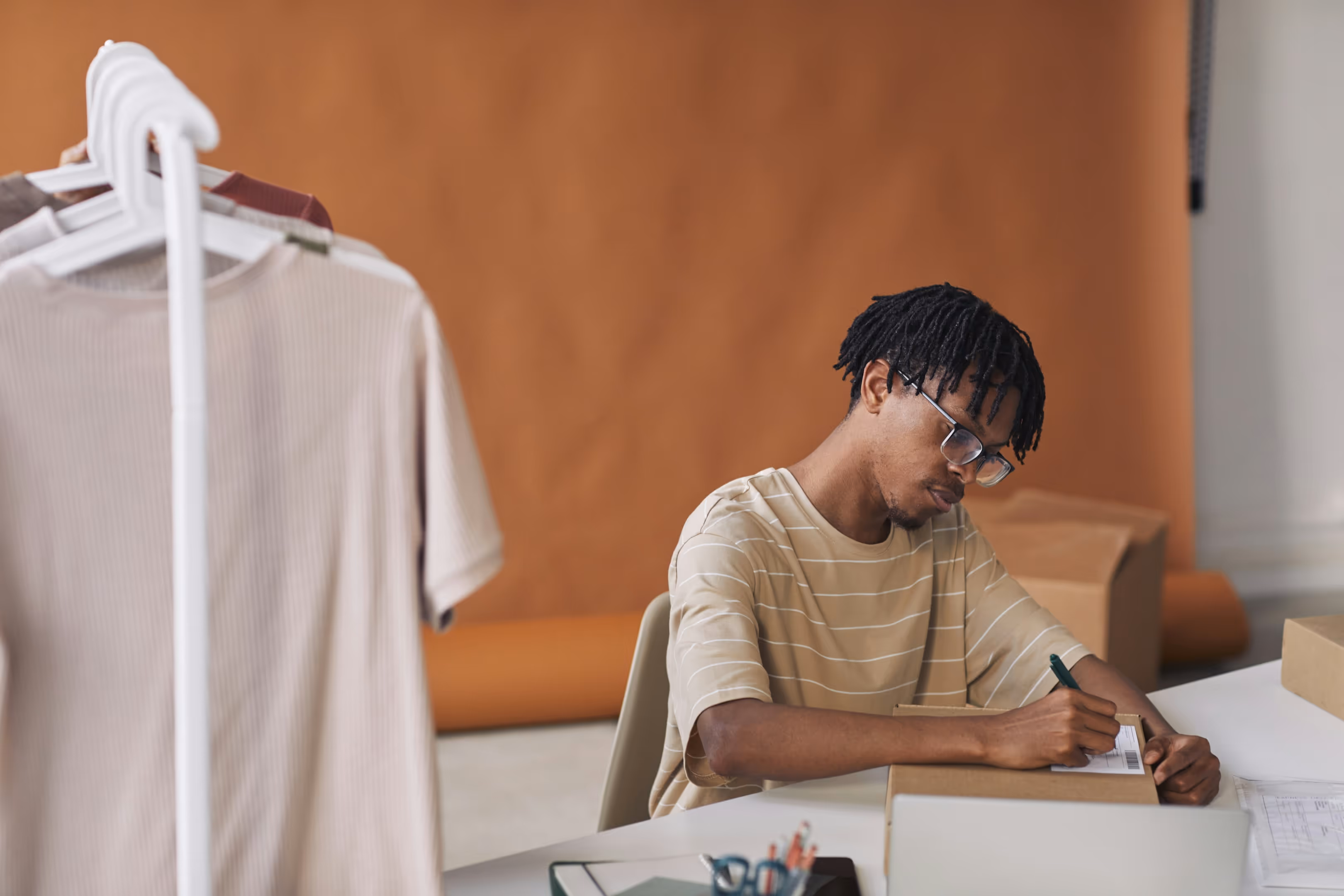 A clothing business owner writing on a box label in his office with a clothing rack behind him.