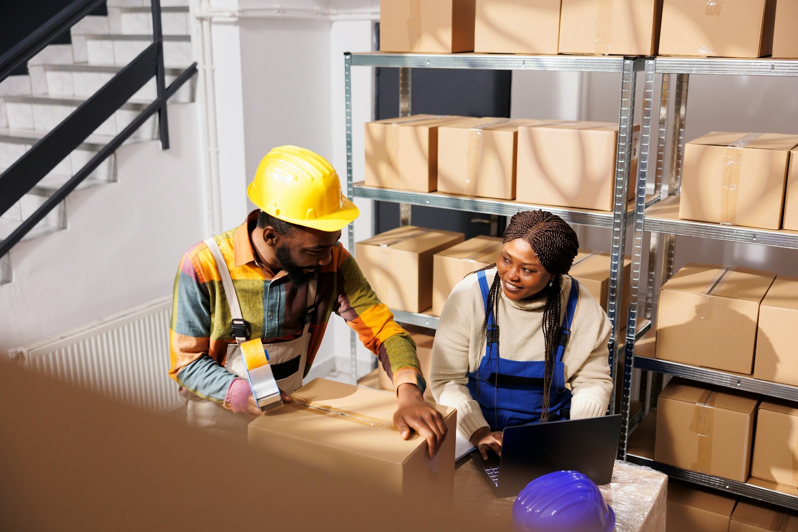 Two warehouse workers: one taping boxes and the other looking at a clipboard and training the worker.