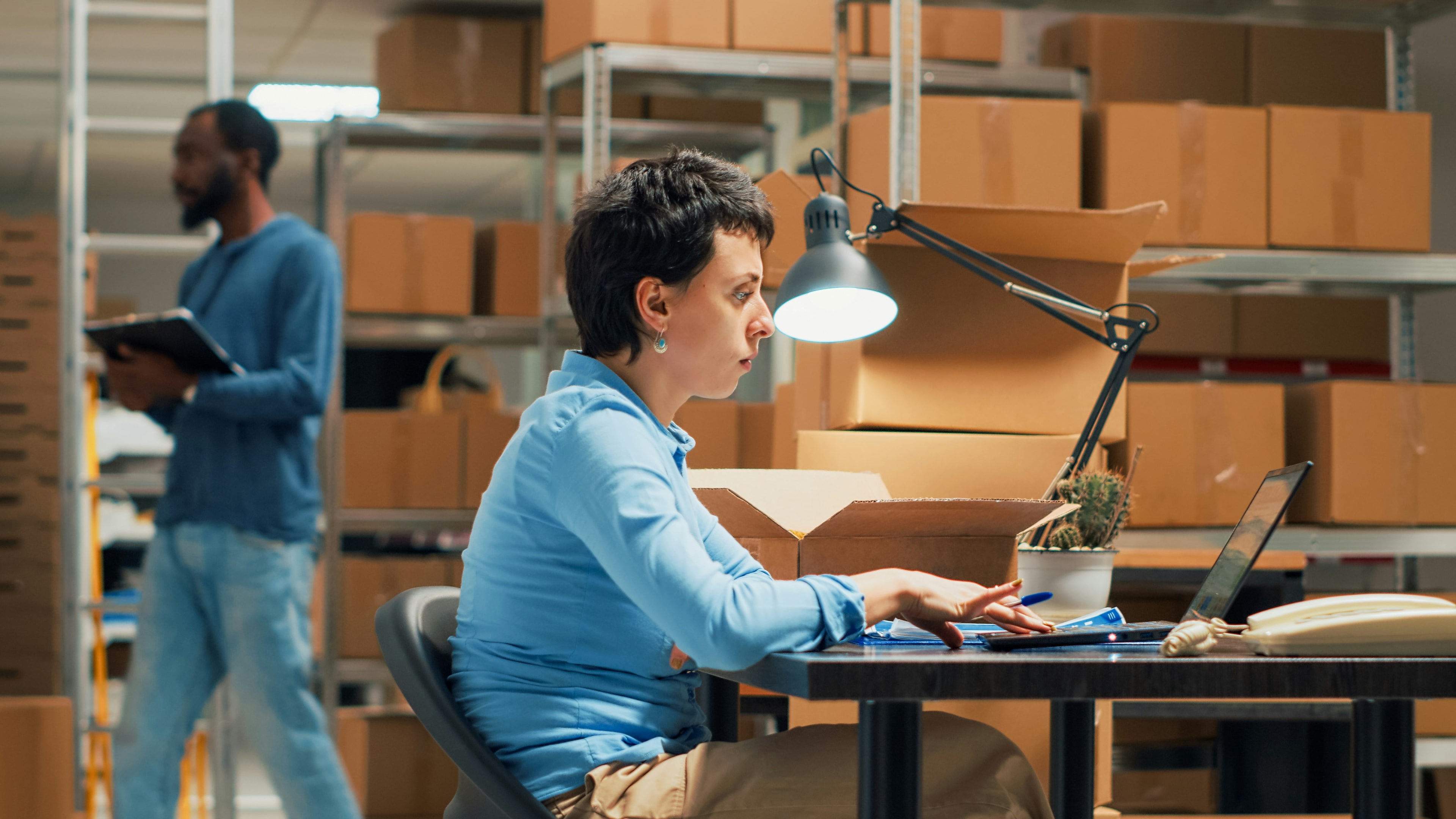 A woman sitting at a desk surrounded by shelves of boxes while a worker checks inventory in the background.