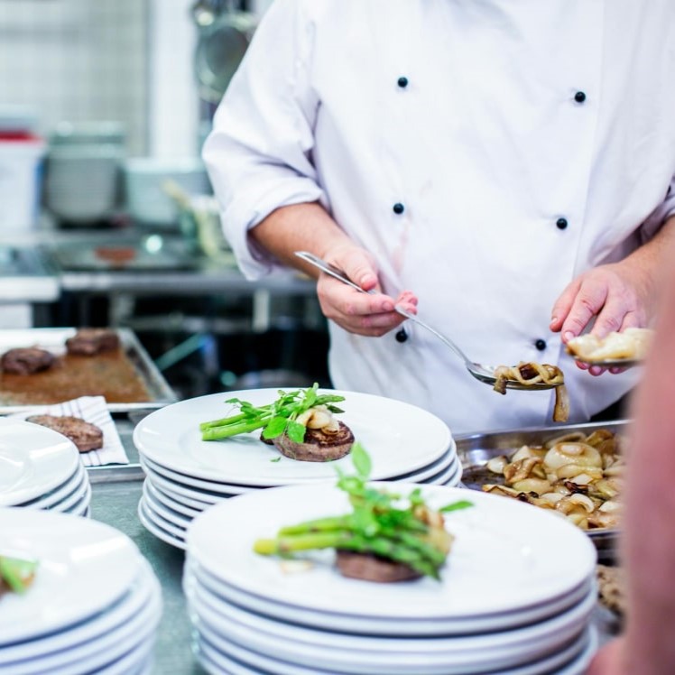 A chef in a white jacket plating meals beside stacks of white dinner plates.