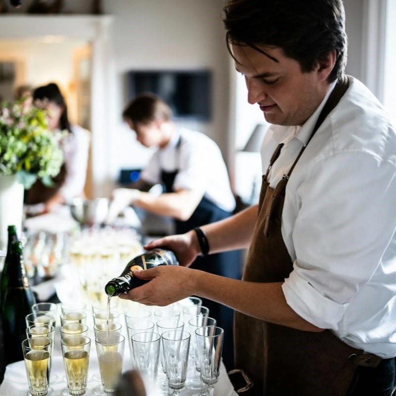 A server pouring sparkling wine into glasses arranged on a service table.