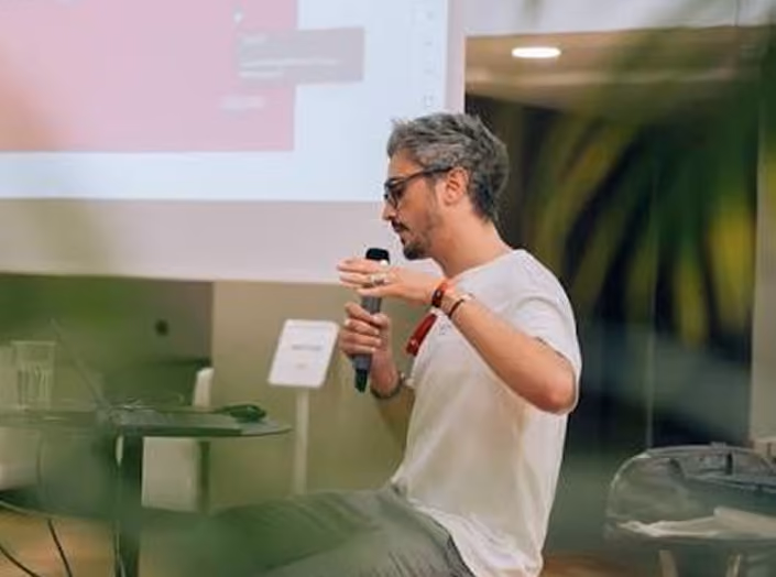 Tanguy ALQUIER parlant dans un microphone lors d'une présentation en intérieur avec un écran en arrière-plan.