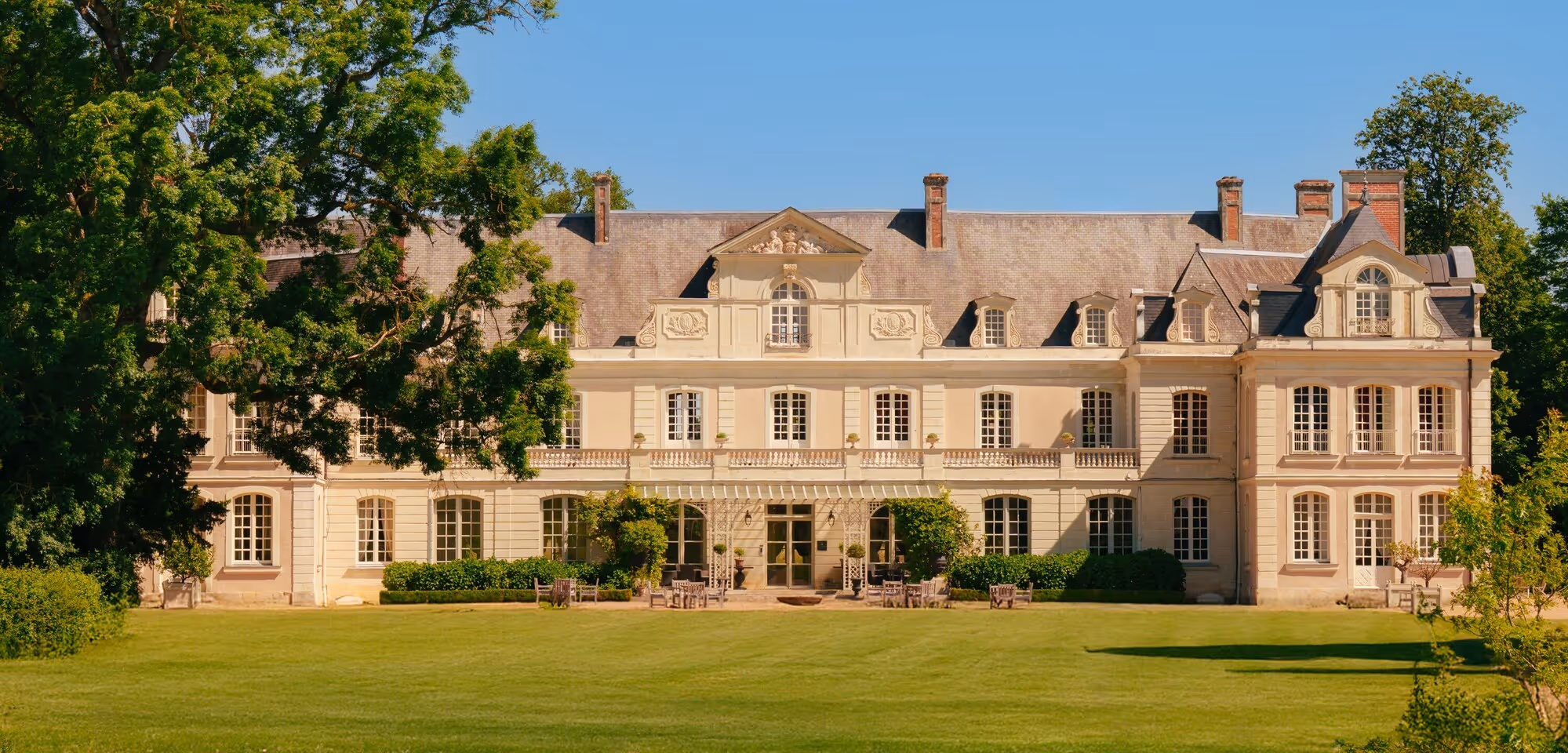 Vue extérieure d'un grand château classique avec un toit en ardoise, de nombreuses fenêtres symétriques, entouré d'arbres et d'une pelouse bien entretenue sous un ciel bleu clair.