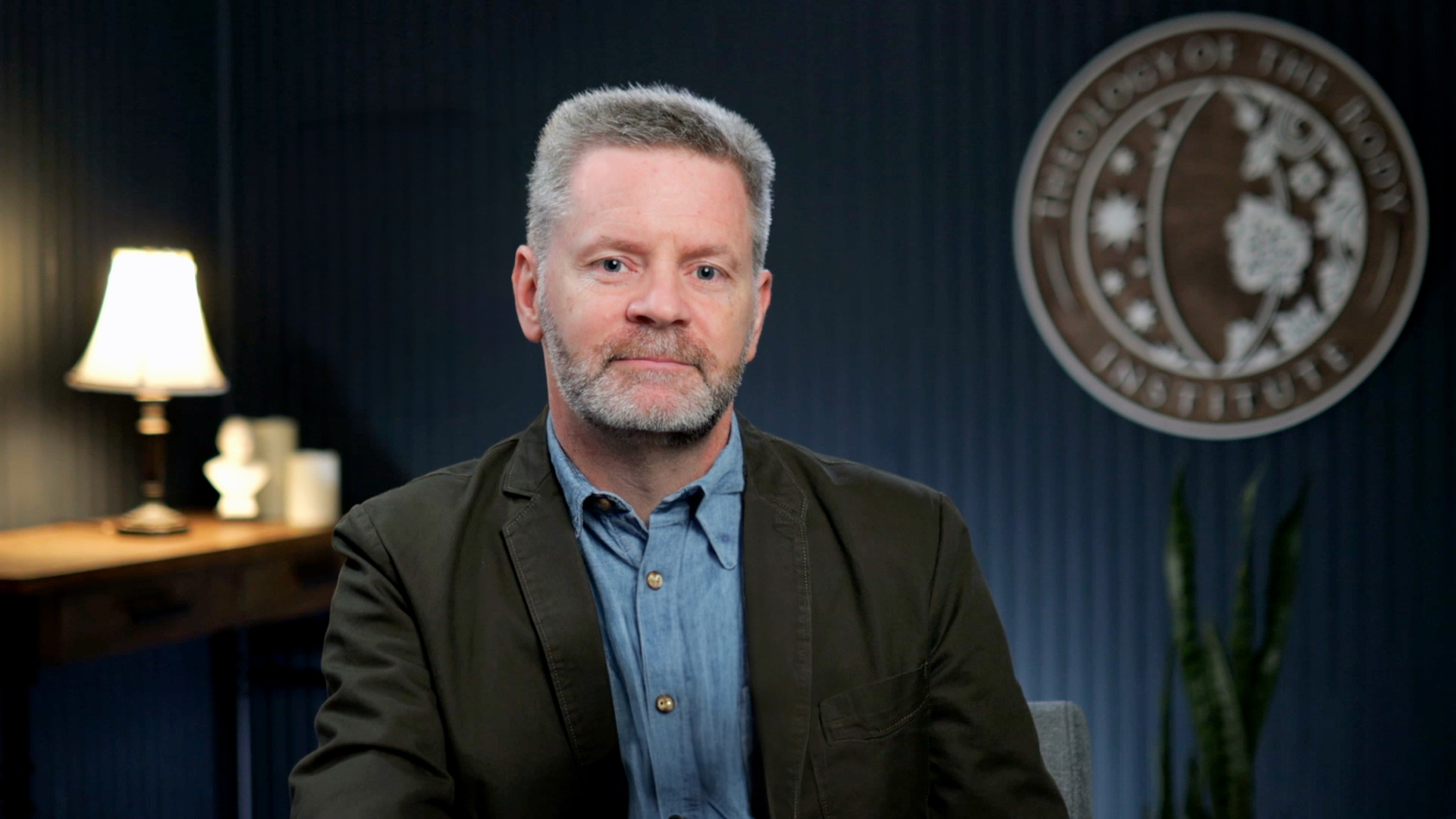 Christopher West: Middle-aged man with gray hair and beard wearing a dark jacket over a blue shirt sitting indoors with a lit lamp and a decorative emblem on a dark wall behind him.