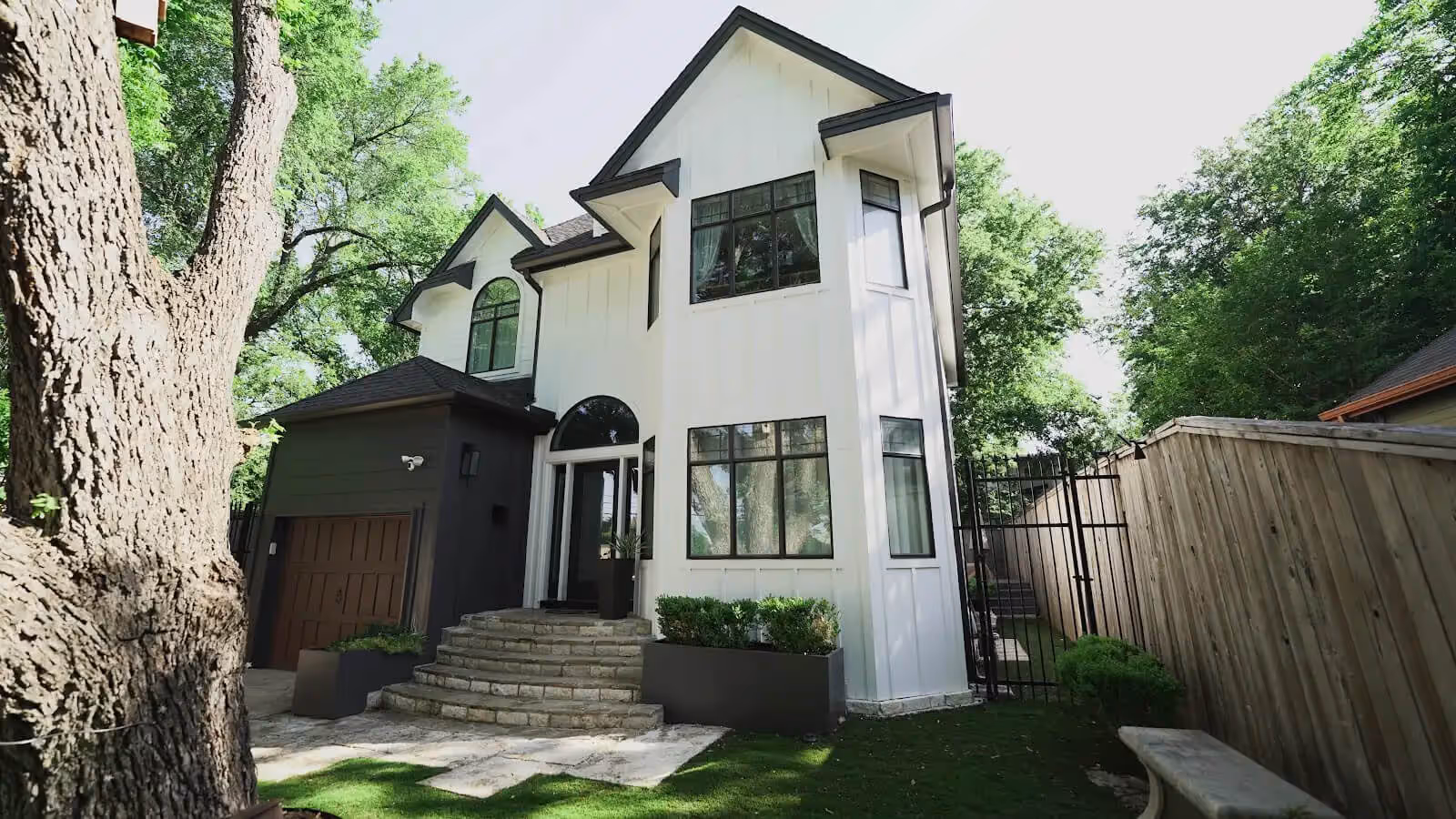 A modern two-story house with a mix of white and dark exterior walls. The front features large windows, including a prominent arched window above the entrance. Stone steps lead up to the front door, which is flanked by decorative plants. On the left, a large tree adds greenery, and a fenced yard is visible on the right, with a pathway and a bench. The setting is surrounded by lush foliage, conveying a serene outdoor atmosphere.
