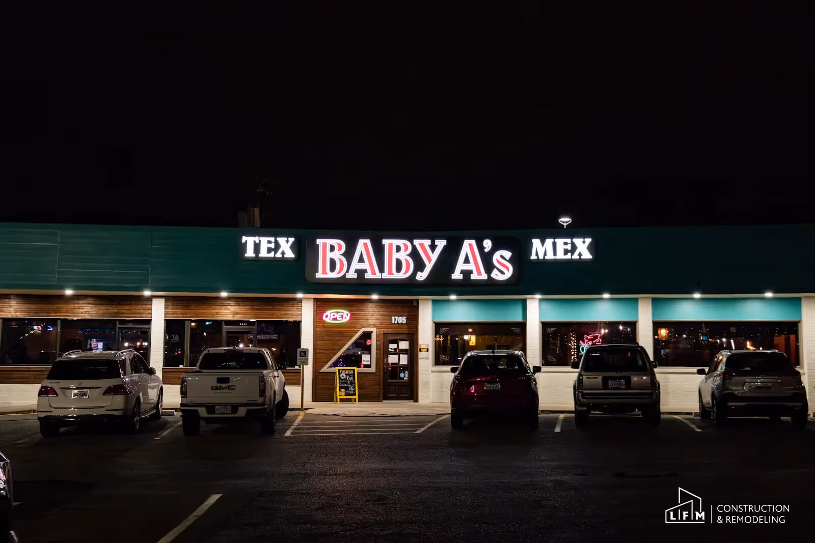 Exterior view of a restaurant called Baby A's, which features a green and wood-paneled facade illuminated by bright lights. The name "BABY A's" is prominently displayed in large, white letters, with "TEX" and "MEX" in smaller text on either side. Beneath the sign, there is an illuminated "OPEN" sign in red. A small chalkboard with a menu or special is visible next to the front entrance. The parking lot is filled with several vehicles, including a white SUV, a grey pickup truck, and other cars, all parked in front of the restaurant. The scene is set at night, highlighting the bright signage against a dark background.