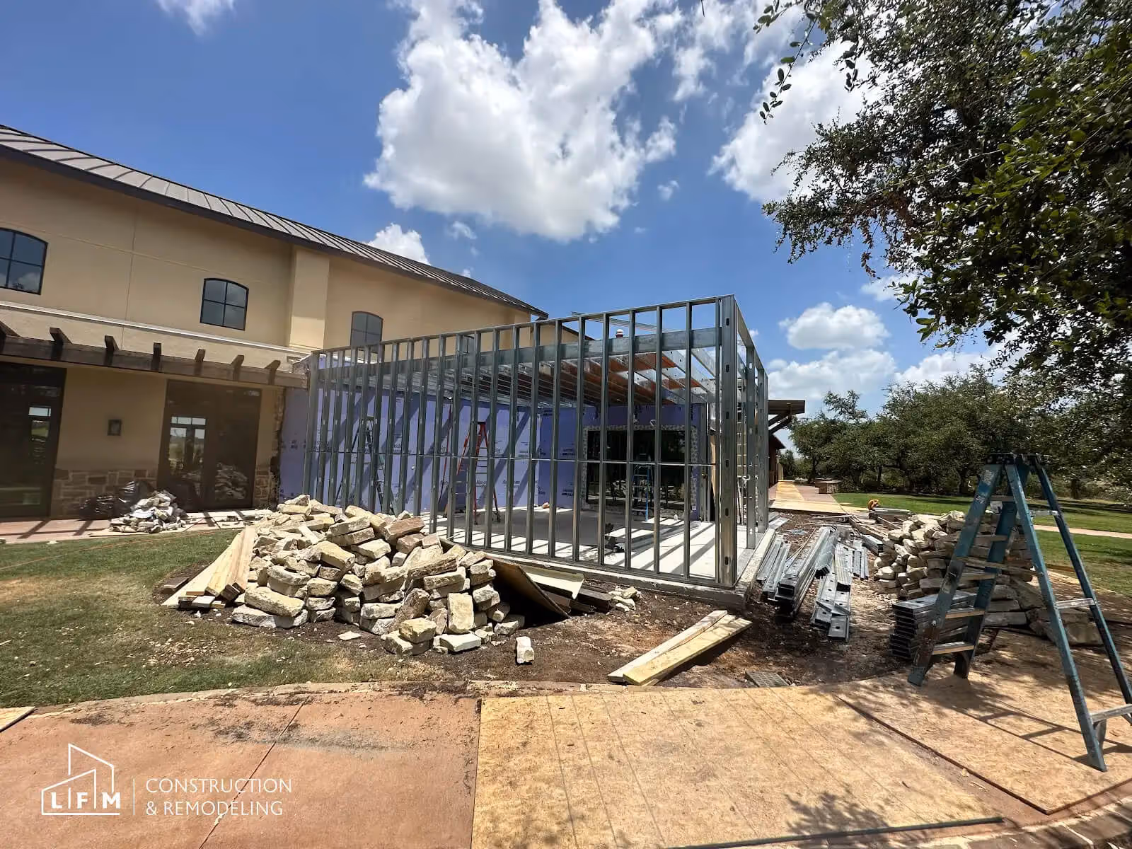 Construction site showing a partially built structure with metal framing on the right, attached to a larger building in the background. The surrounding area is cluttered with construction materials, including piles of stone and metal beams. A blue ladder is positioned near the framework. The sky is bright with a few clouds, and there are trees visible on the right side. The ground is a mix of dirt and paved surfaces. The building features large windows and a shaded patio area.