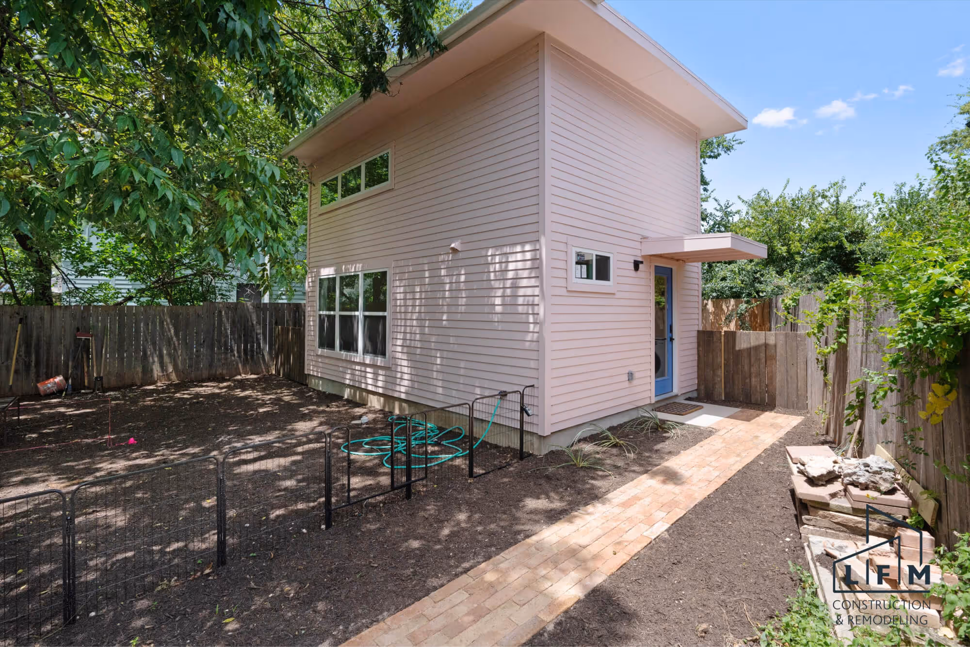 Exterior view of a modern, two-story house with light pink siding, featuring large windows. The front door, painted blue, is located to the right, leading to a small patio area with a welcome mat. A narrow brick pathway extends from the door, bordered by freshly mulched garden soil. Along the left side, there is a black metal fence enclosing a yard that appears to have been recently cleared, with a green garden hose coiled nearby. Lush green trees provide shade above, and a wooden fence encloses the property in the background. The scene is bright with blue skies visible above.