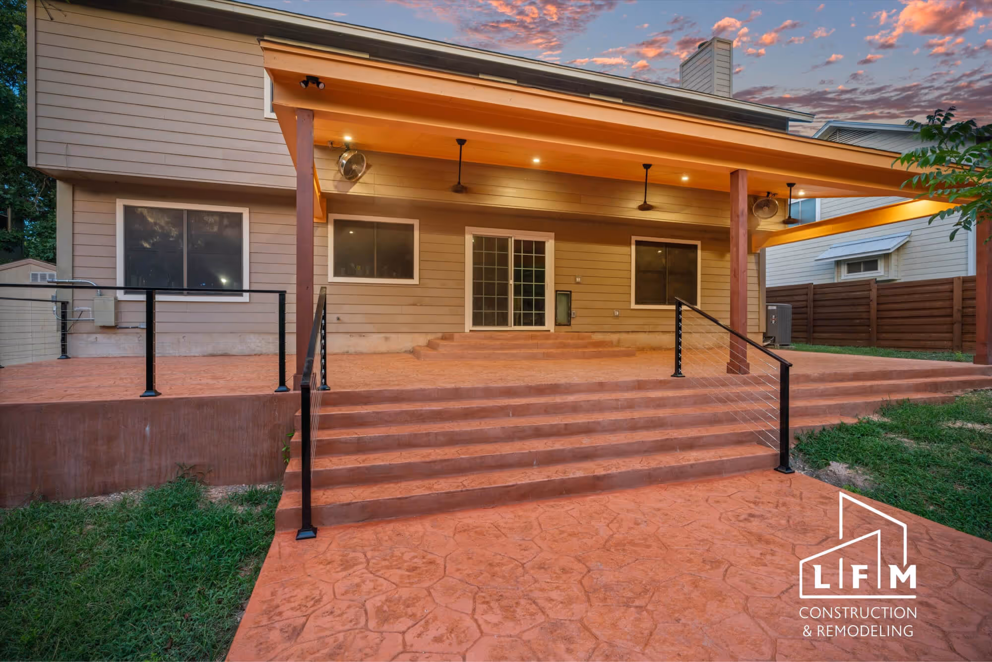 A backyard patio area featuring a set of stone-patterned concrete steps leading up to a sliding glass door. The steps are flanked by black metal railings. Above the patio, there is an overhanging roof with ceiling fans. The exterior walls of the house are tan, and large windows line the side. The ground is covered with grass, and the space appears well-lit, indicating twilight with colorful clouds in the sky. A logo for LFM Construction & Remodeling is displayed in the bottom right corner.