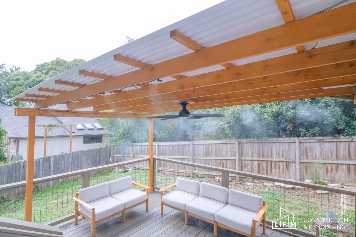 A covered patio area featuring a wooden frame and a metal roof, with a ceiling fan hanging from the beams. There are two sleek, light gray sofas with wooden frames positioned around the space. In the background, a green lawn is visible, bordered by a wooden fence. The scene is bright and airy, with trees providing a natural backdrop.
