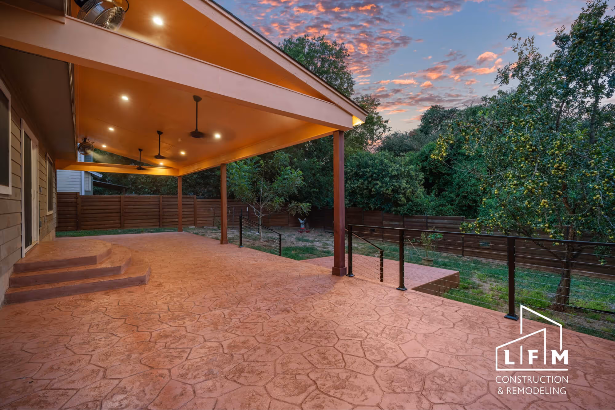 A spacious outdoor patio with a textured, reddish-brown concrete floor, featuring decorative patterns. The patio is covered by an orange overhang with recessed lighting and several ceiling fans. To the left, there are three steps leading up to the interior of a house with vertical wooden siding. In the background, lush green trees and a wooden fence are visible, partially obscuring the yard. The sky is filled with soft pink and purple clouds, indicating sunset. The bottom right corner includes a logo that reads "LFM Construction & Remodeling."