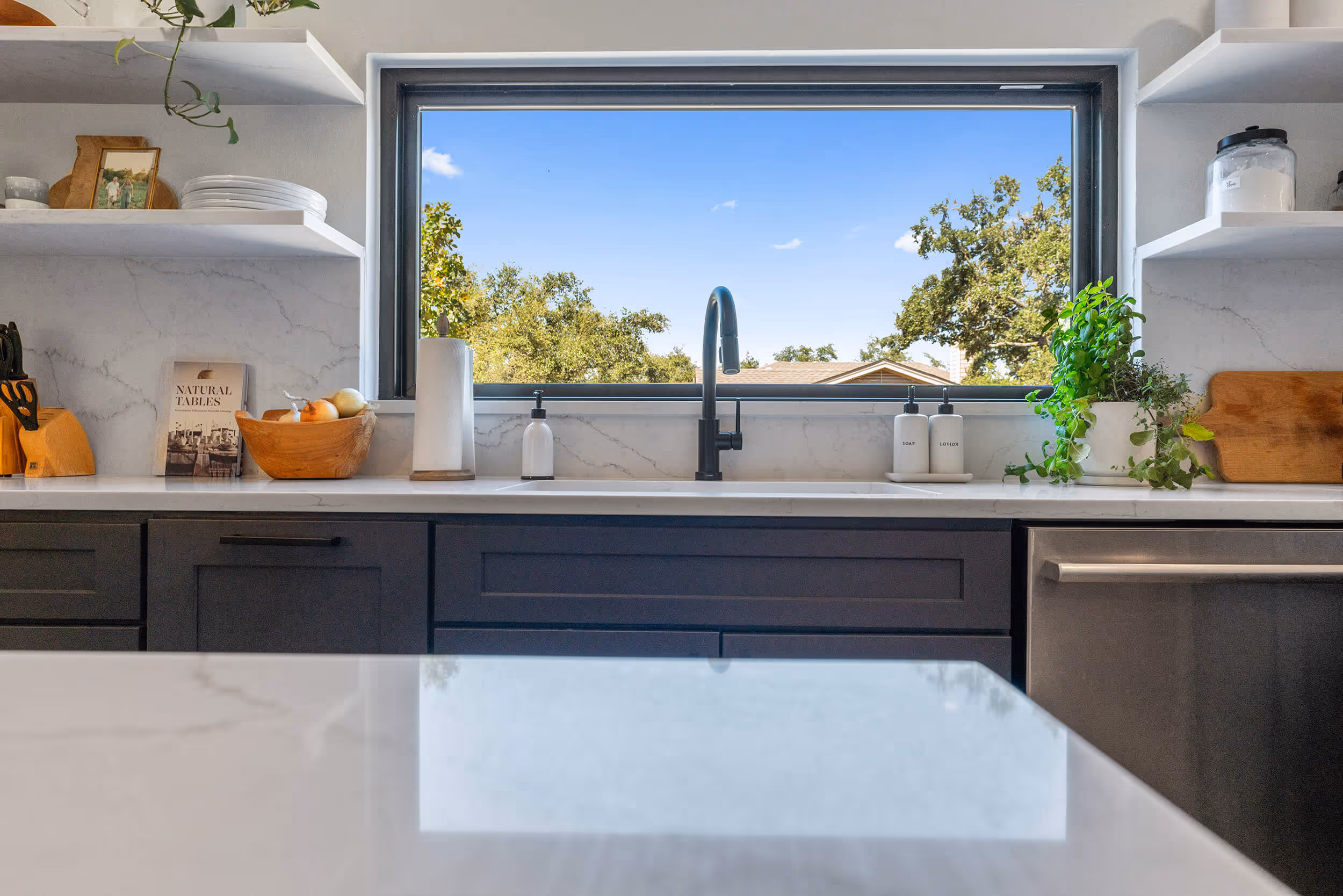 A modern kitchen scene featuring a view through a large window. The window frame is dark and lets in bright sunlight, illuminating a marble countertop in the foreground. The countertop has a slight reflection and is mostly clear of items. In the background, there are two floating shelves with various items including a small wooden bowl of apples, a cookbook titled "Natural Tables," kitchen utensils, and two white soap dispensers. Green plant life is visible near the window, adding a touch of nature to the kitchen. The view outside includes trees and a clear blue sky dotted with a few clouds.