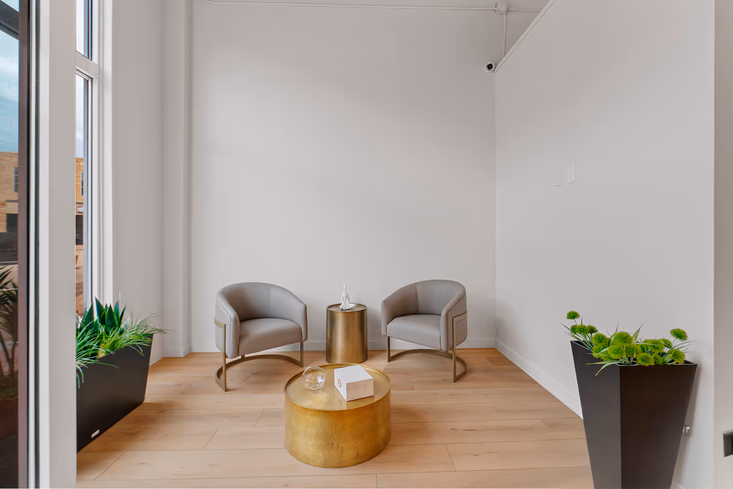Modern waiting area with two gray chairs, round gold tables, and green potted plants on wooden flooring.