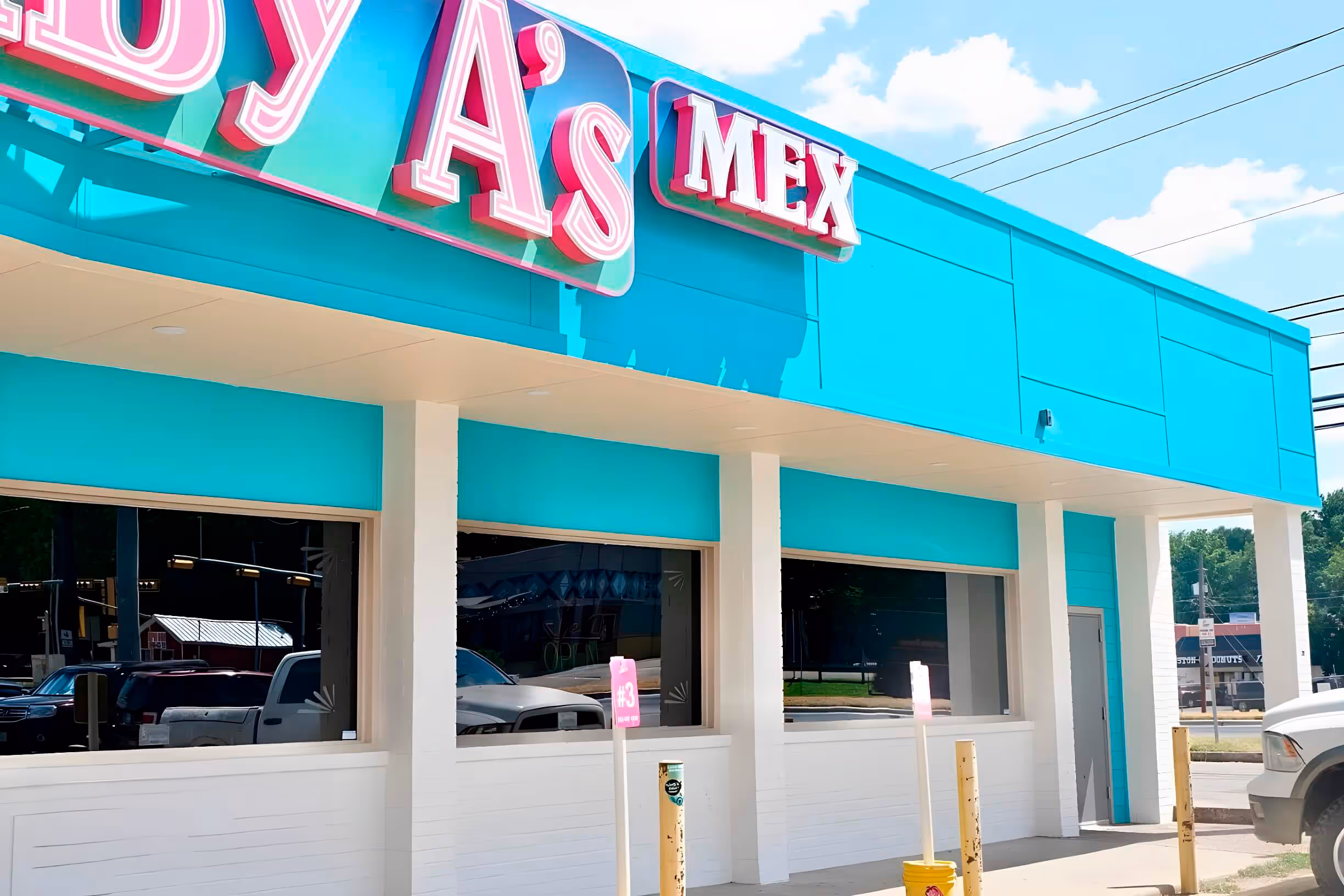 Bright turquoise building with a pink neon sign, featuring large windows and parking spaces in front.