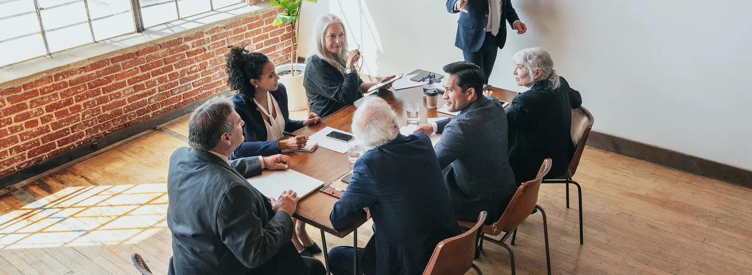 Business meeting with six professionals seated around a wooden table in a bright office with a brick wall and large windows.
