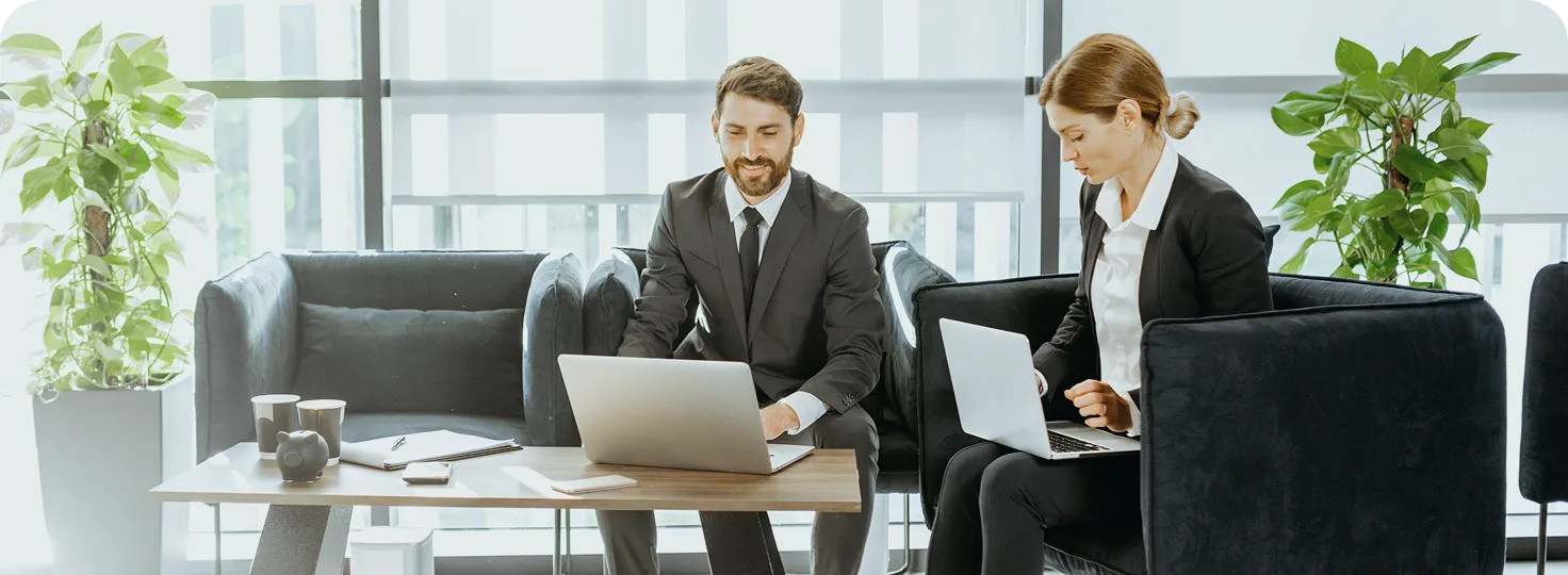 Two business professionals in suits working on laptops while seated in a modern office lounge with plants and a wooden coffee table.