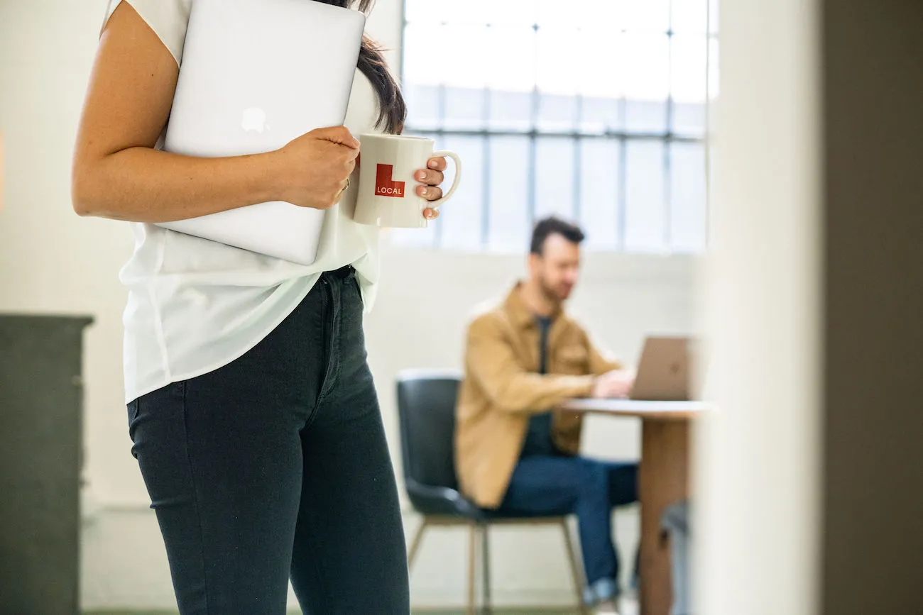 Lady carries Local-branded mug and laptop through office as man works from wooden table under large industrial window in the background