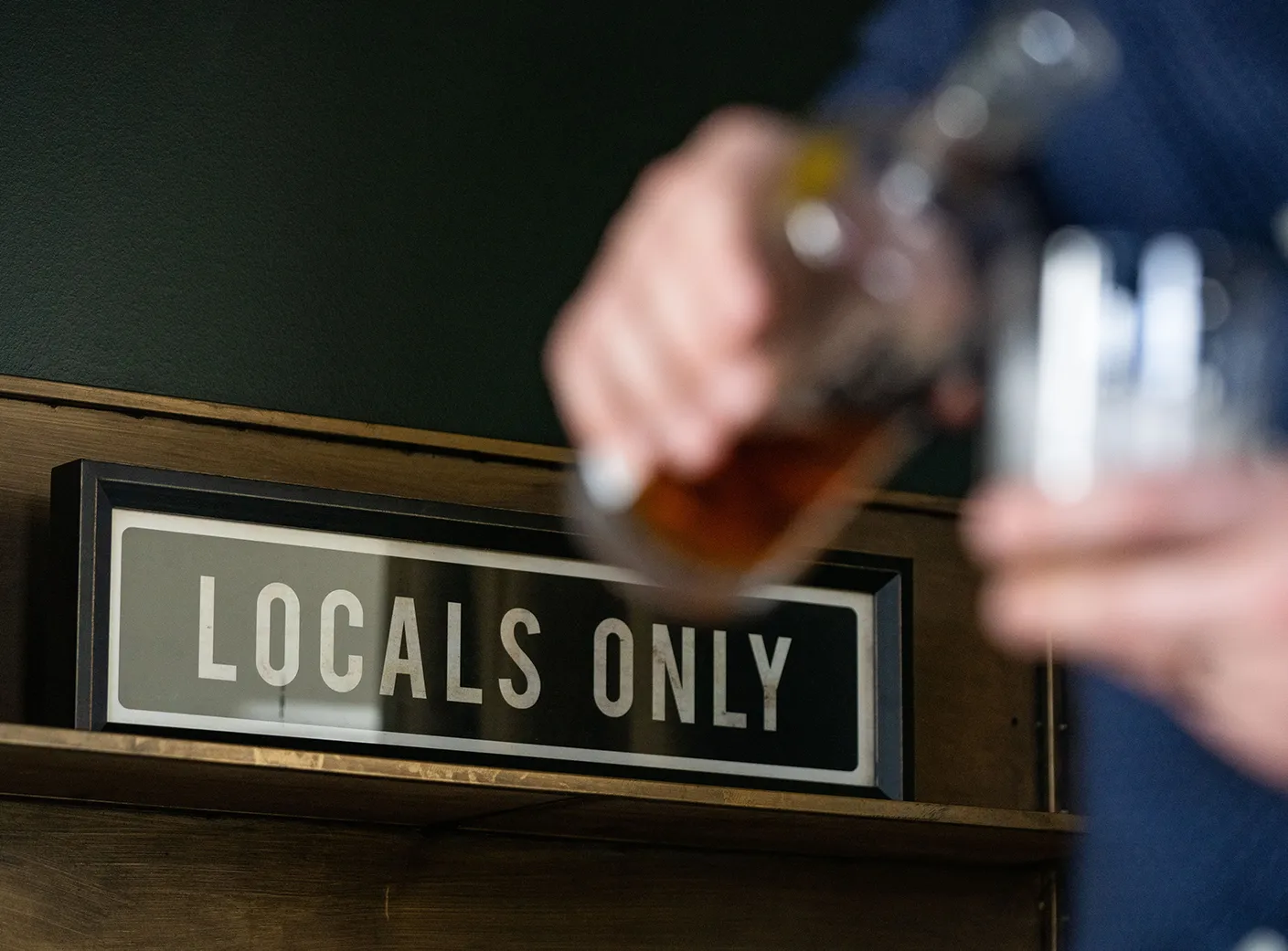 Man pours glass of bourbon in front of "Locals Only" sign