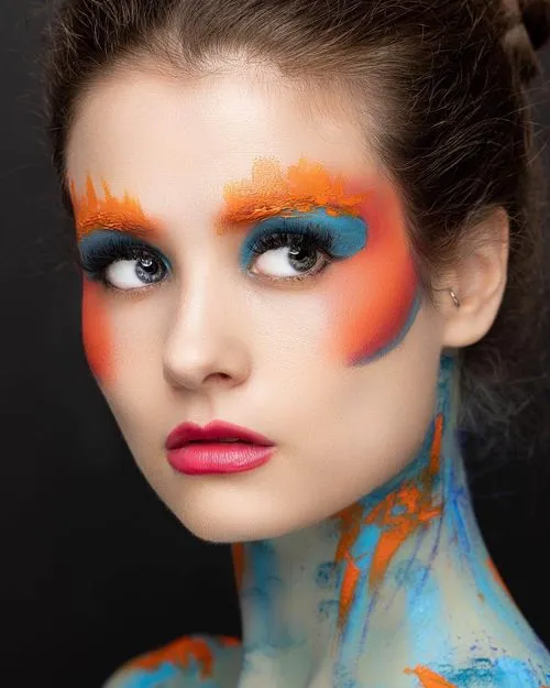 Woman with vibrant blue and orange makeup and abstract paint smeared on her neck.