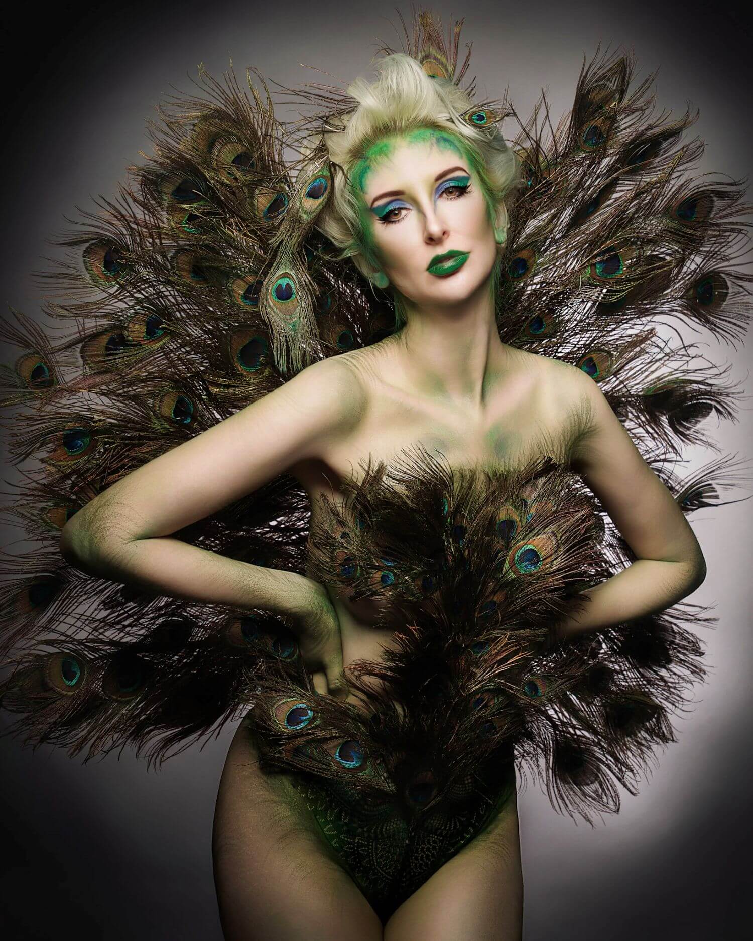 Woman in green full body painting and peacock feathers, hands on hips, by OKOS Creations.