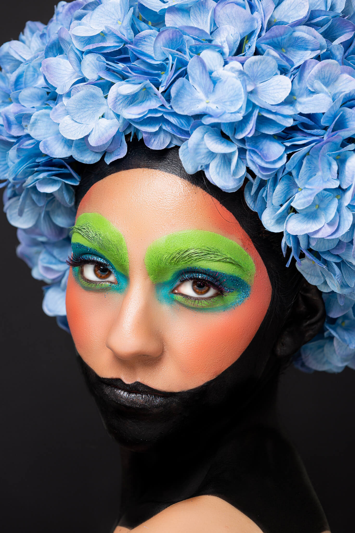 Woman with bold green painted eyes, donning a blue floral afro, makeup designed by OKOS Creations.