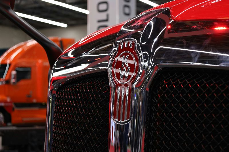 Close-up of a red Kenworth semi truck grille and emblem inside a modern diesel truck repair and maintenance facility.