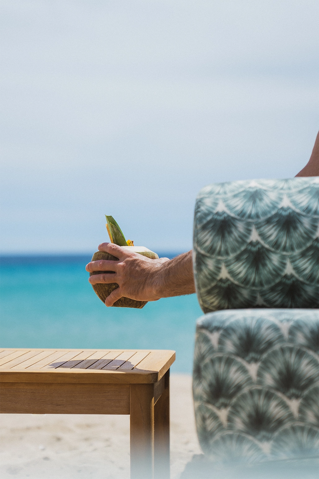 Main tenant un coco fraîchement coupé devant une chaise aux motifs verts sur une plage avec une mer turquoise en arrière-plan.