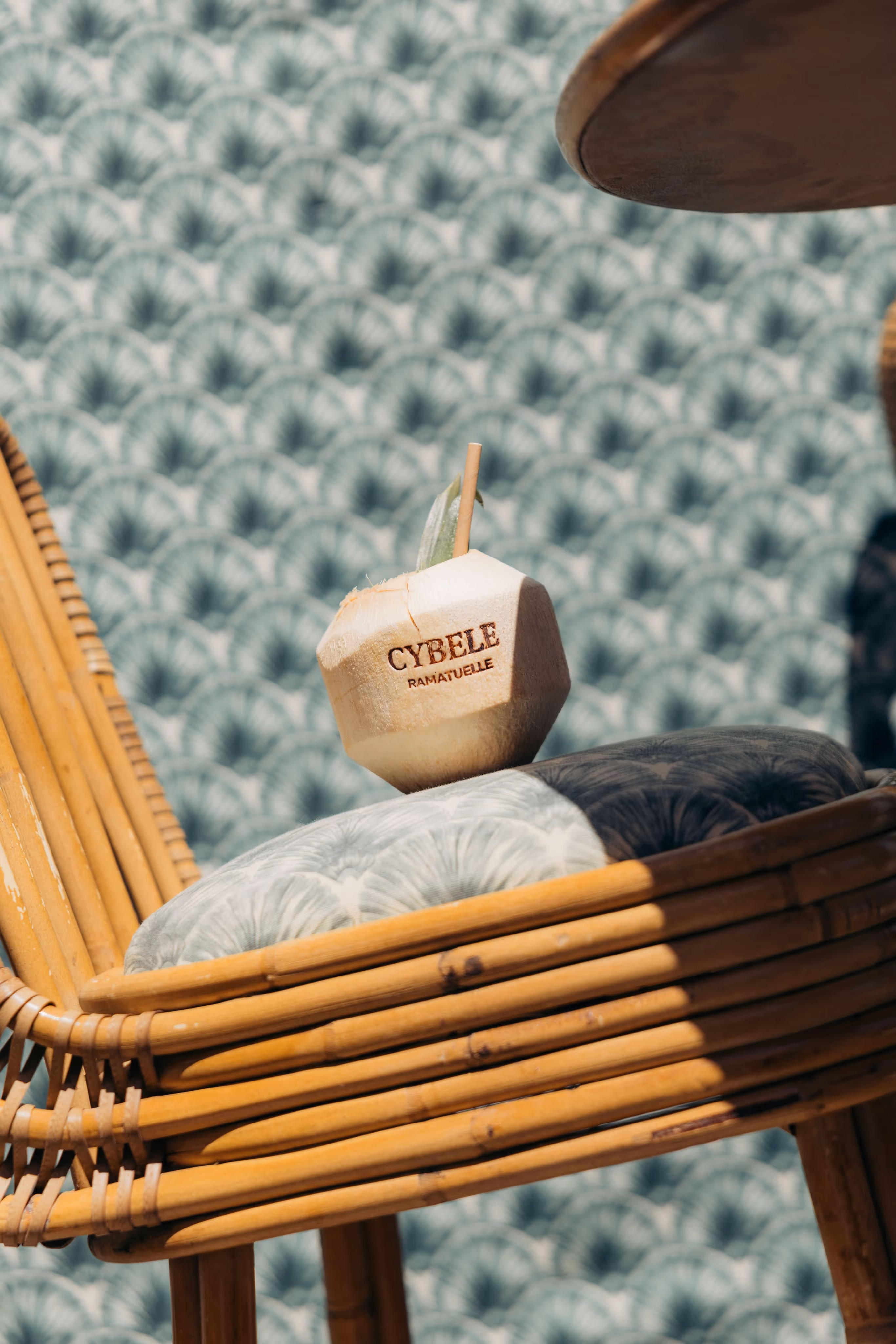 Noix de coco avec paille portant l’inscription Cybele posée sur un coussin et une chaise en rotin.