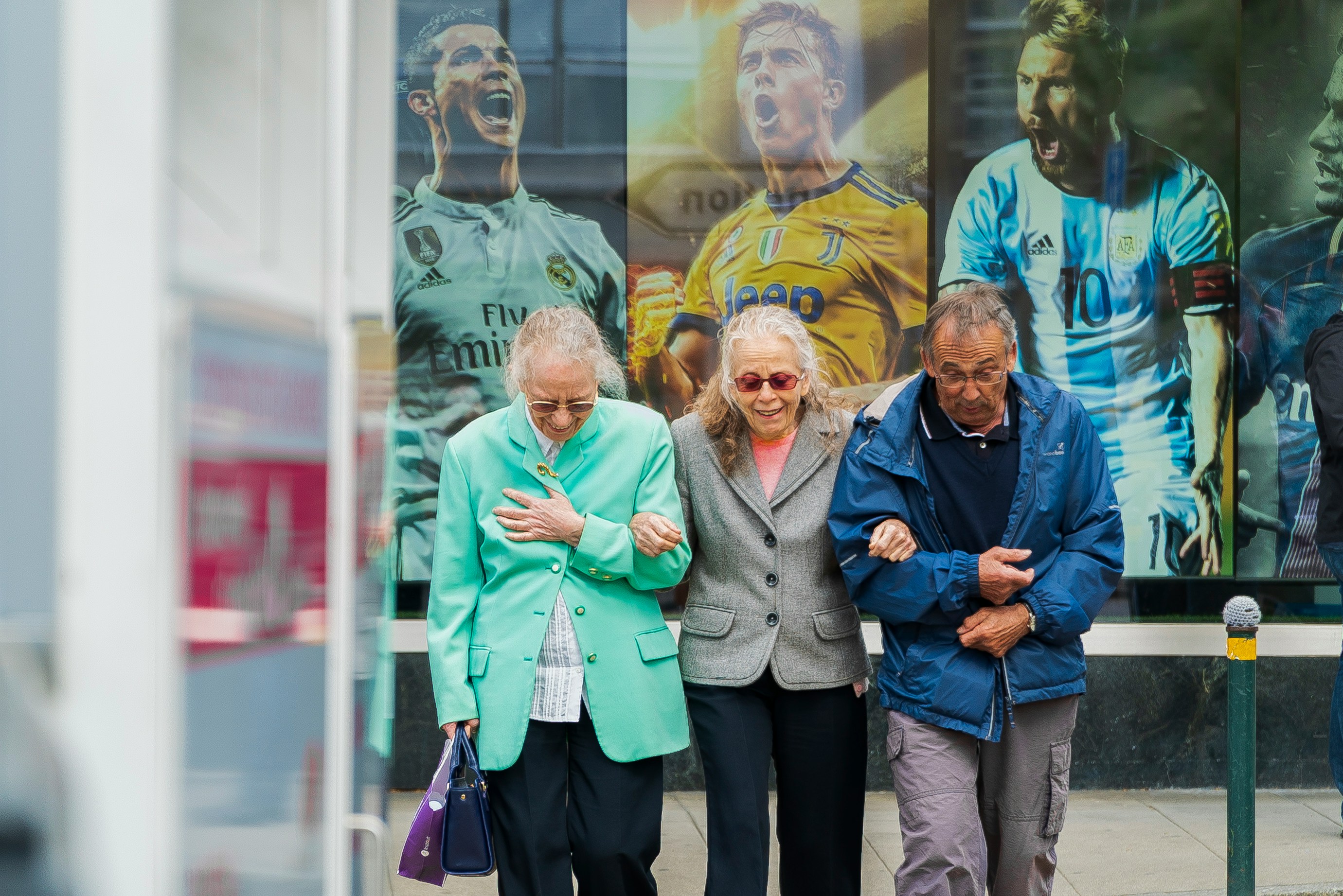 The image depicts three elderly individuals walking arm-in-arm on a city street, displaying warmth and mutual support. In the background, a large advertisement features famous soccer players in team jerseys with Adidas branding. The scene contrasts the energy of sports culture with the companionship of the elderly group, emphasizing themes of community, support, and the intersection of everyday life with popular culture.