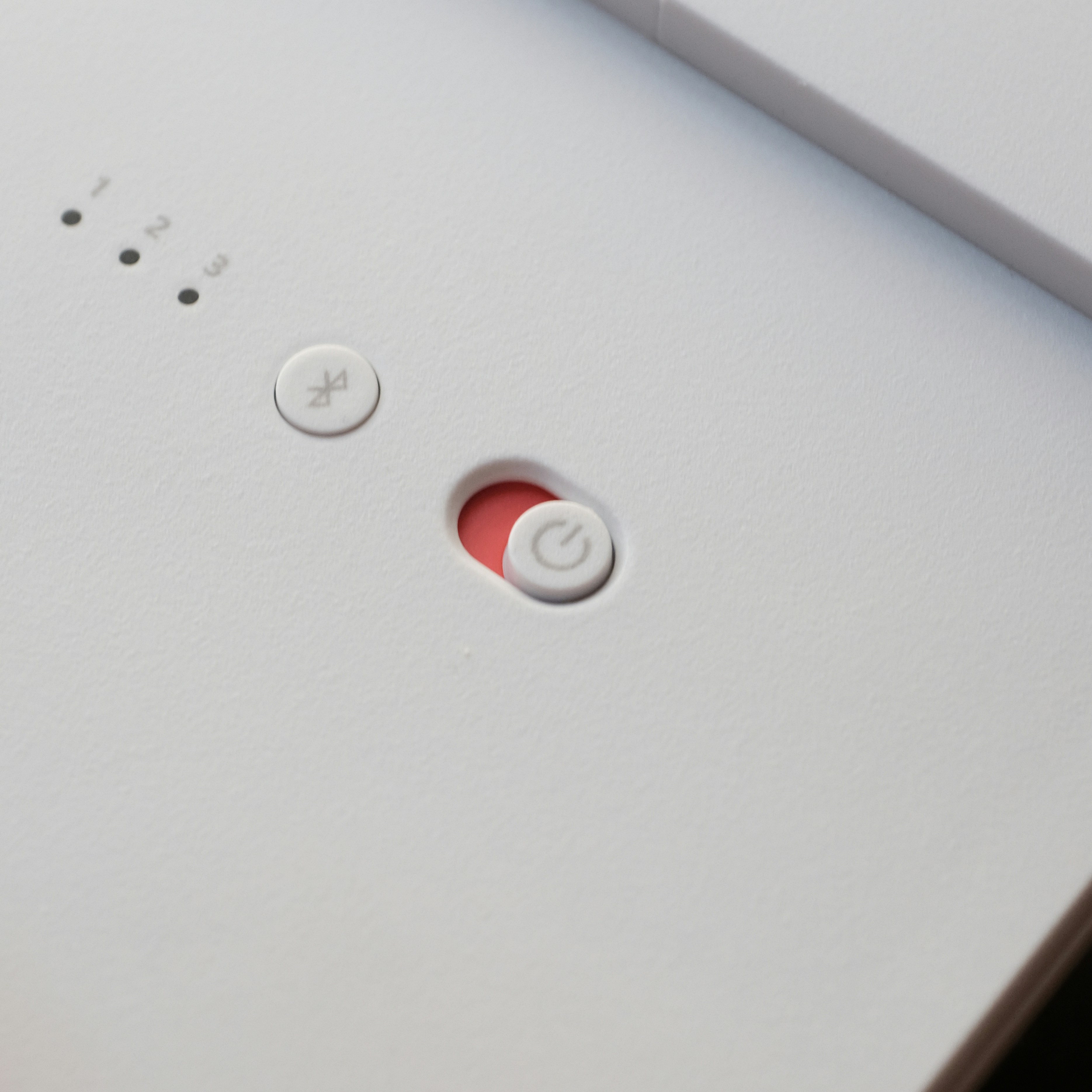The image depicts a close-up of a white electronic device with a minimalist control interface. Key features include a power button, a mute button, and indicator lights labeled with numbers, emphasizing the device's essential operational controls and simple design.
