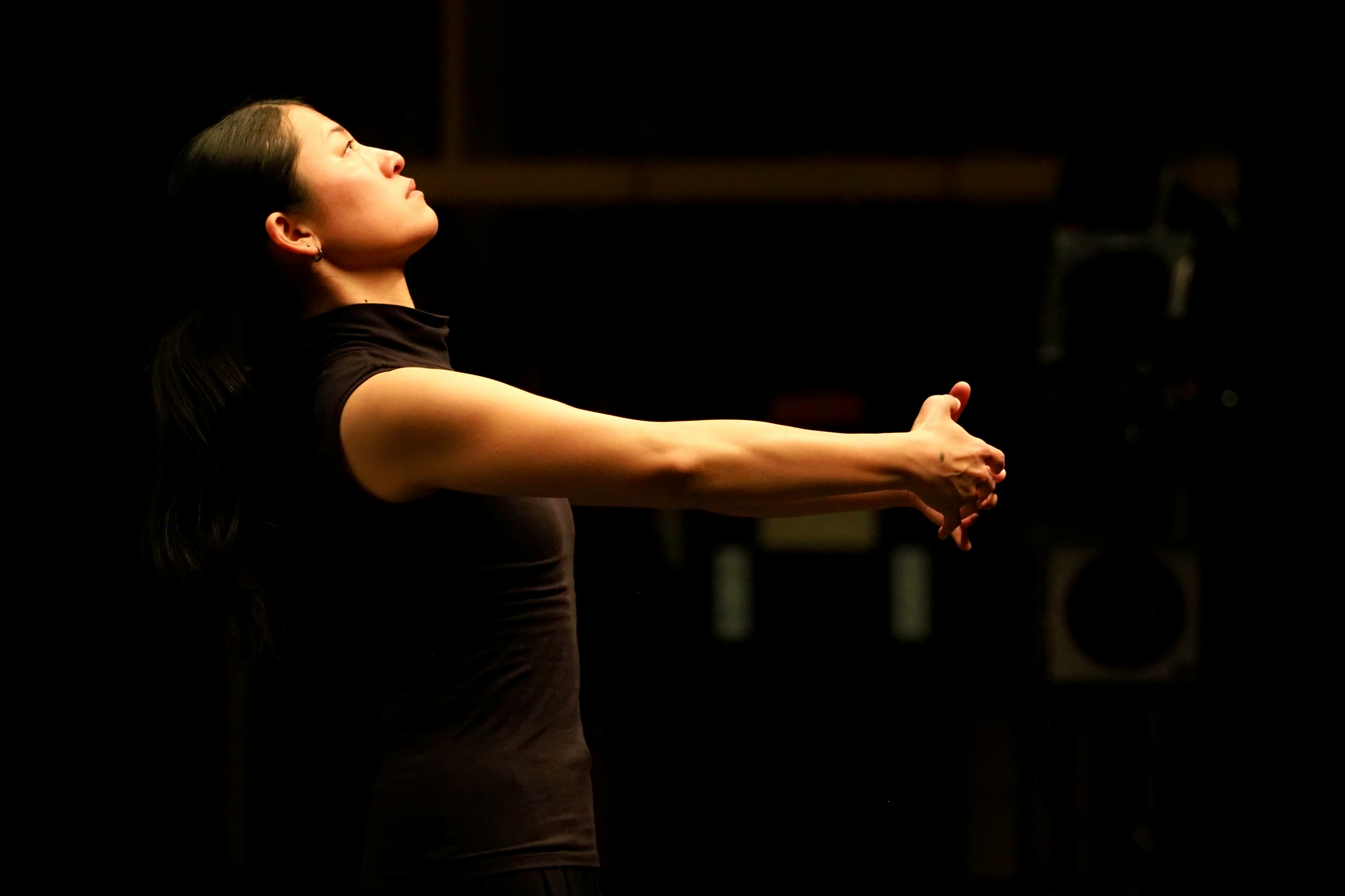 The image depicts a woman in a dark environment, standing with her arms extended forward and her head slightly raised, suggesting she is engaged in stretching, focused concentration, or a performance. The scene conveys themes of mindfulness, preparation, or artistic expression, possibly related to dance, theater, or meditation.